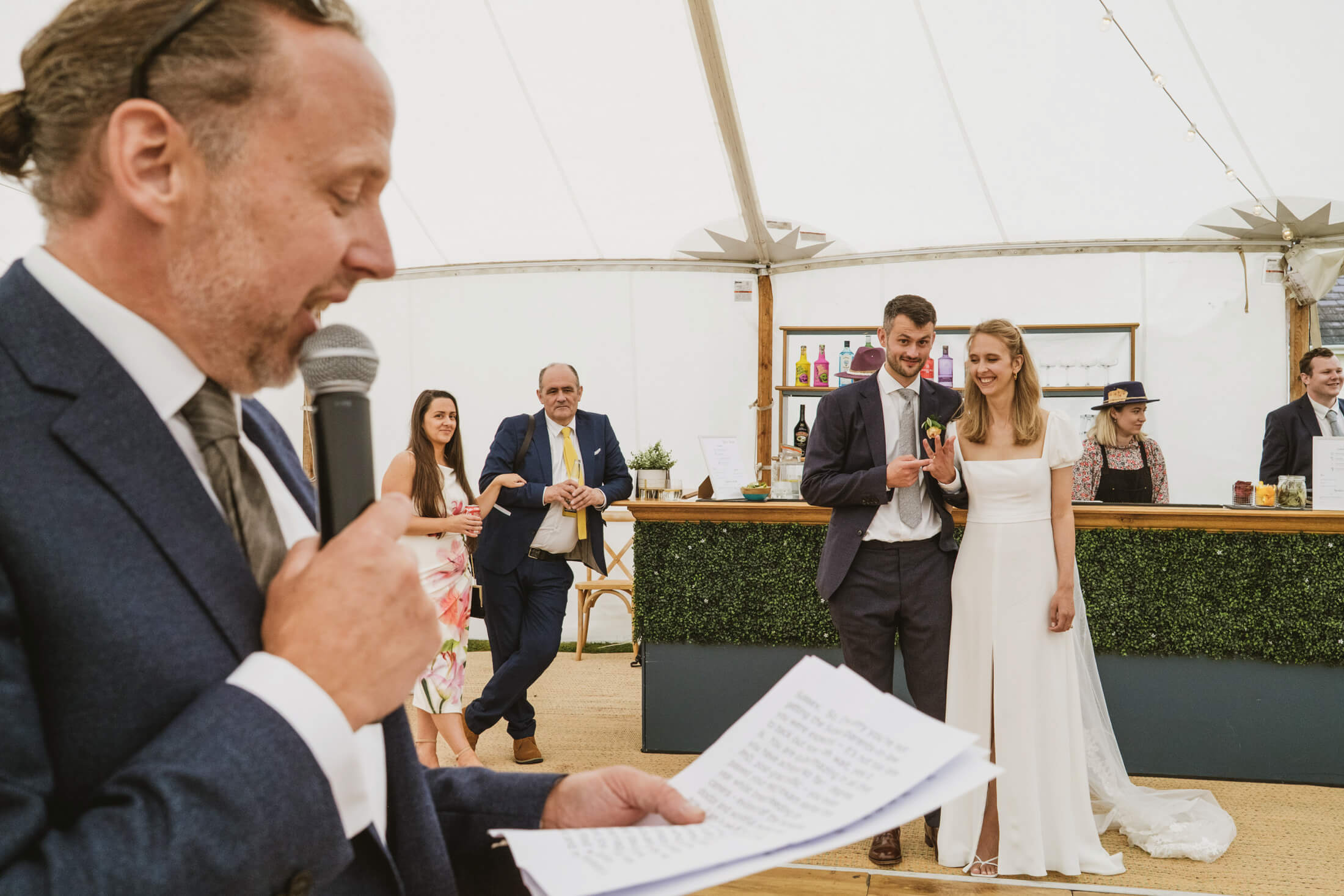 Father of the bride reading speech as couple laugh together at wedding reception bar