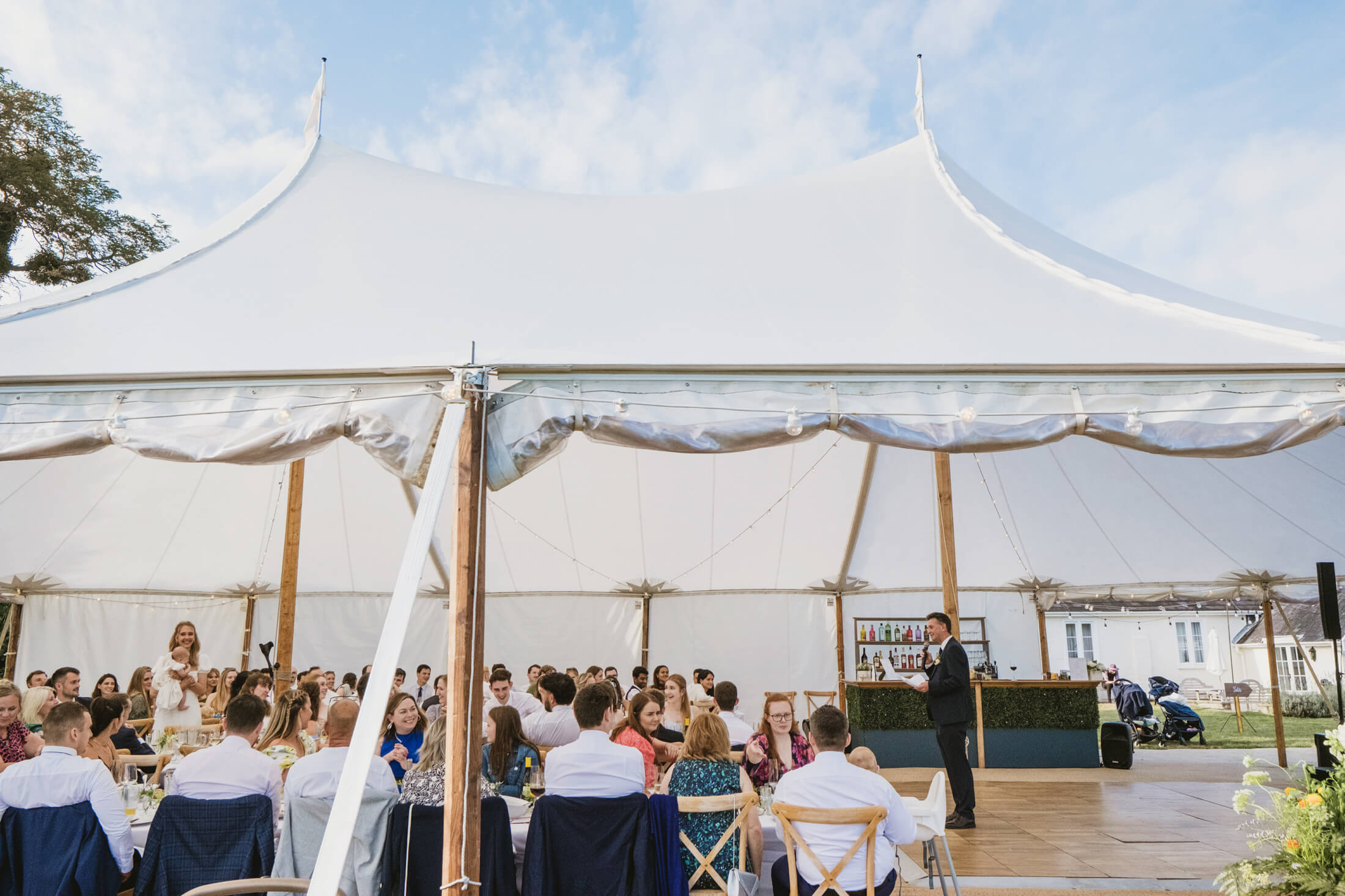 Guests listen to a speech beneath a white marquee at a summer wedding reception.