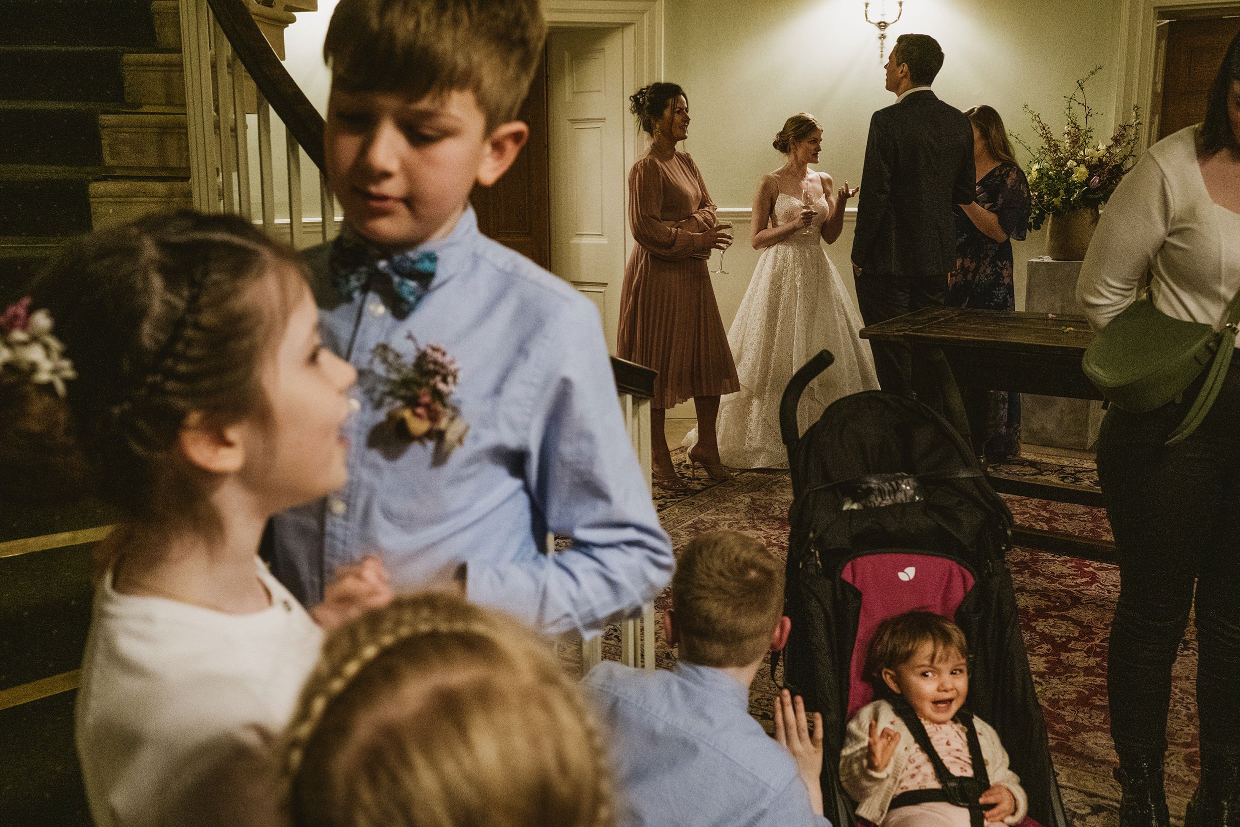 layered photo of bride chatting with guests and children in foreground. Little baby interacting with page boy.