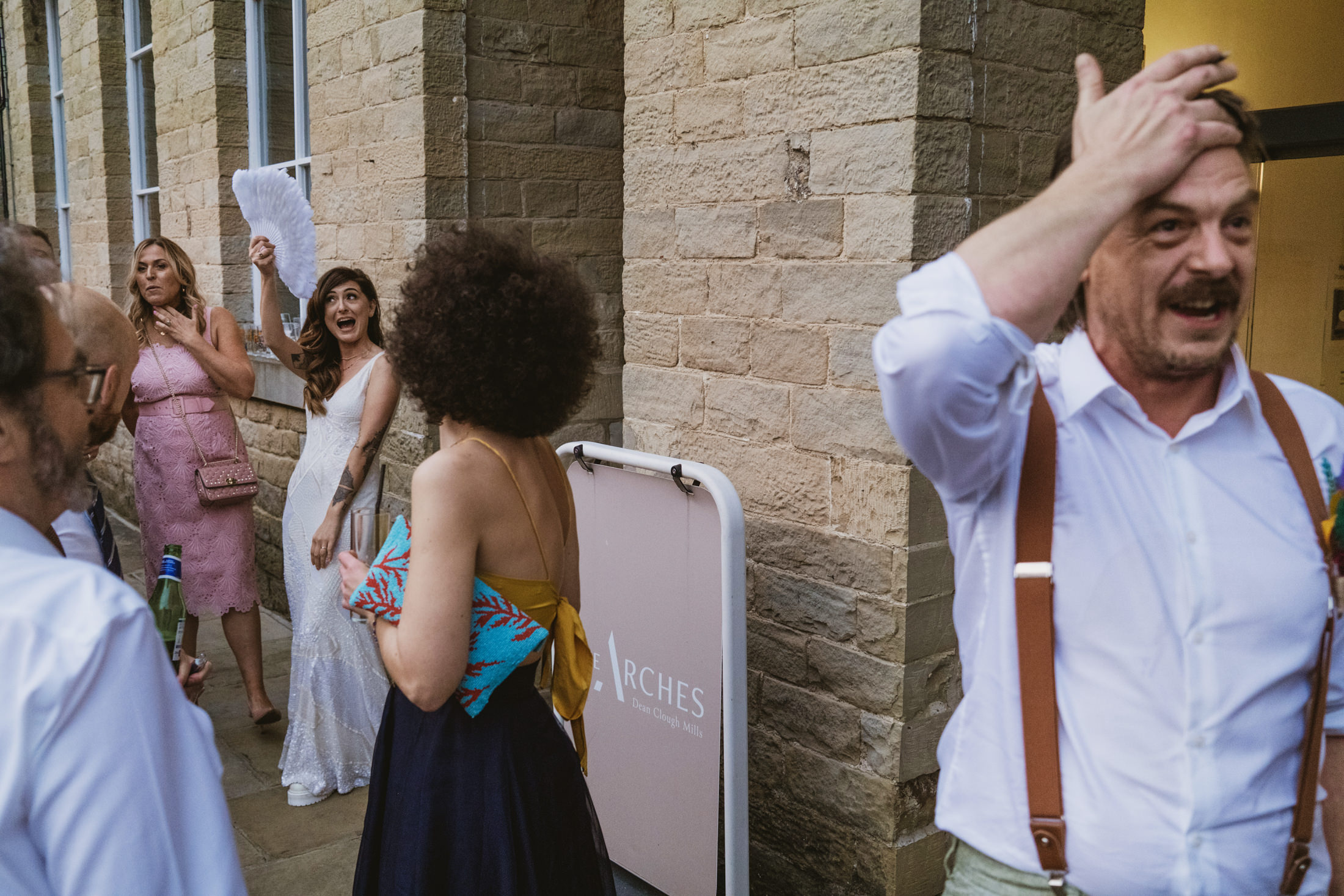 Bride laughing as guest lifts fan outside stone venue, friends reacting in summer light – documentary wedding photography.