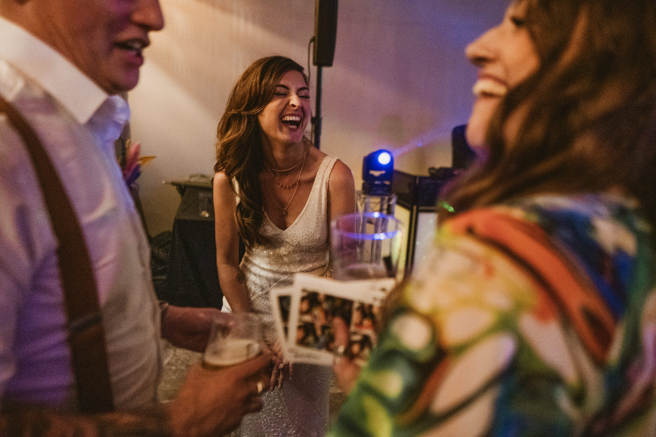 Bride laughing with friends on dance floor at wedding reception, drinks in hand.