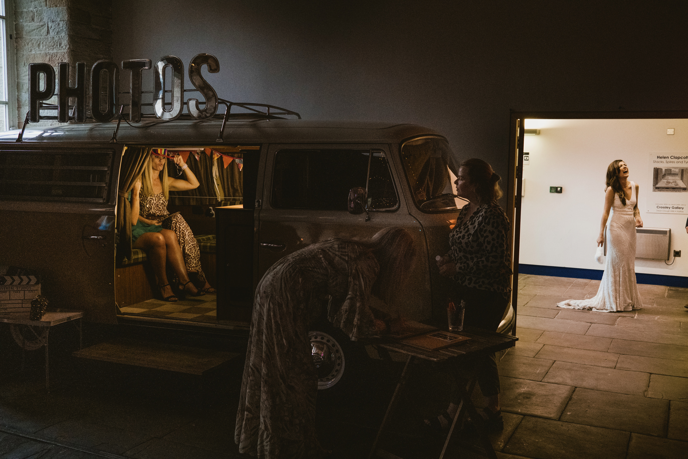 Bride laughing in gallery doorway beside vintage photo booth van at evening wedding reception.