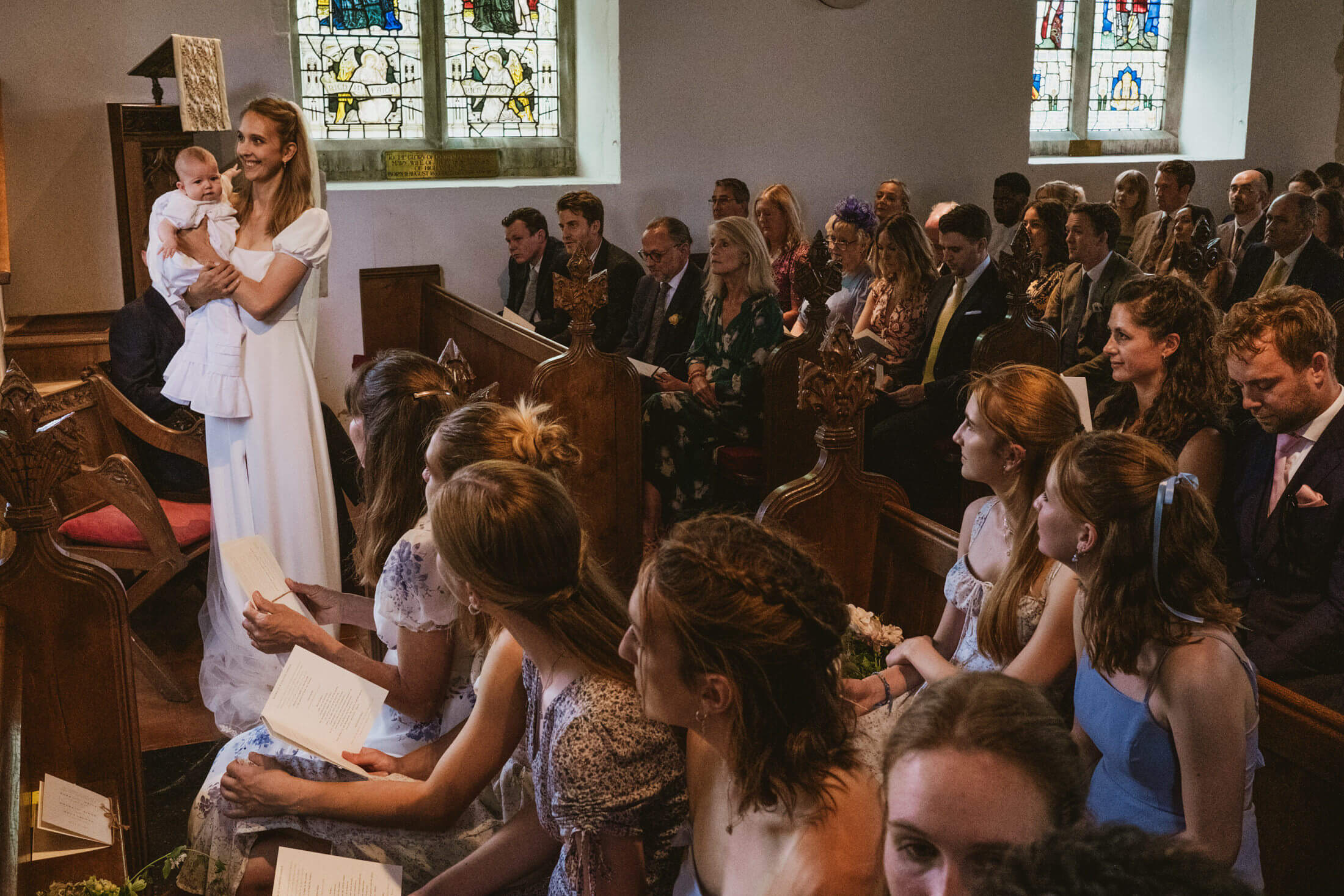 Woman in white dress holds baby in church pews as guests watch, sunlight through stained glass documentary wedding photography.