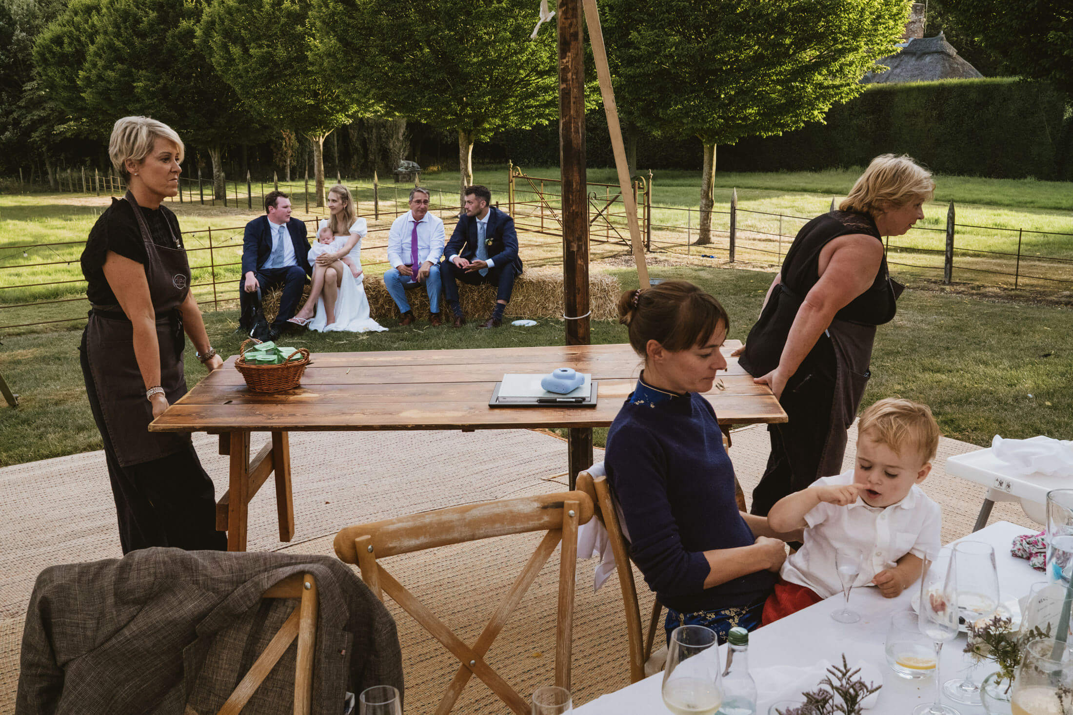 Guests chat on hay bales as a child sits at a table during a garden wedding reception.
