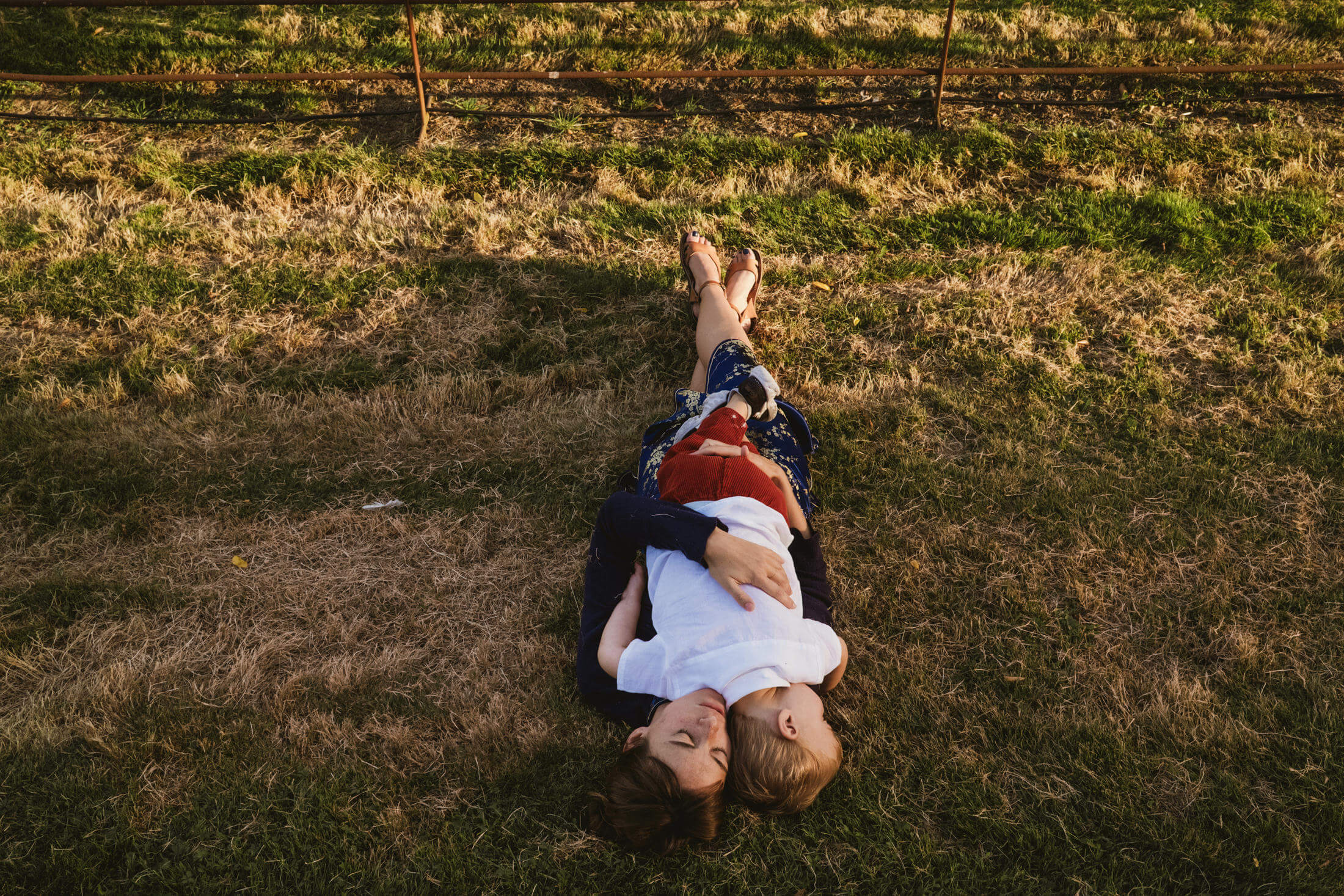 Overhead view of couple lying on sunlit grass, child resting on chest, quiet embrace in golden light wedding photography