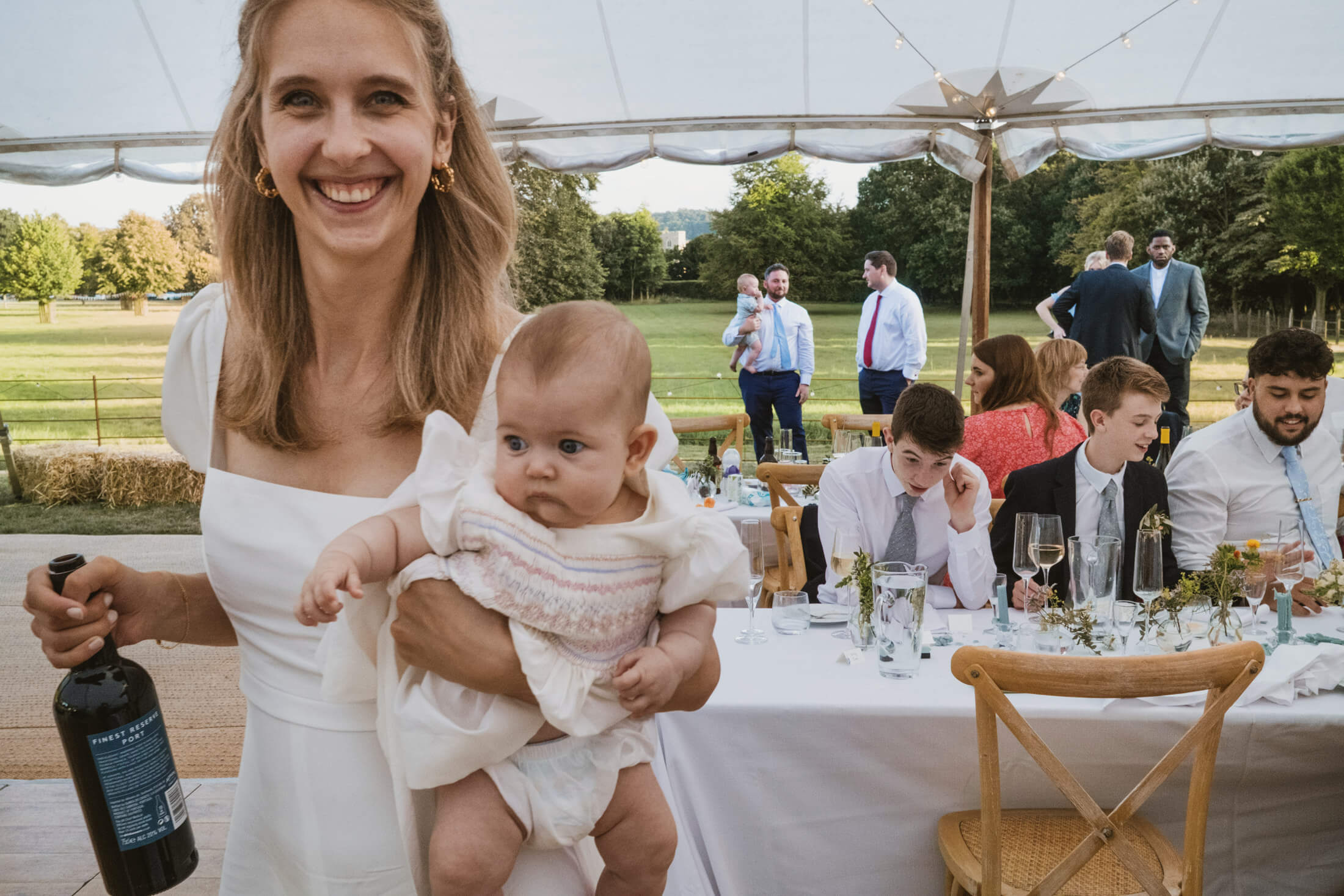 Bride holding baby and wine bottle, smiling at sunlit outdoor wedding reception table.