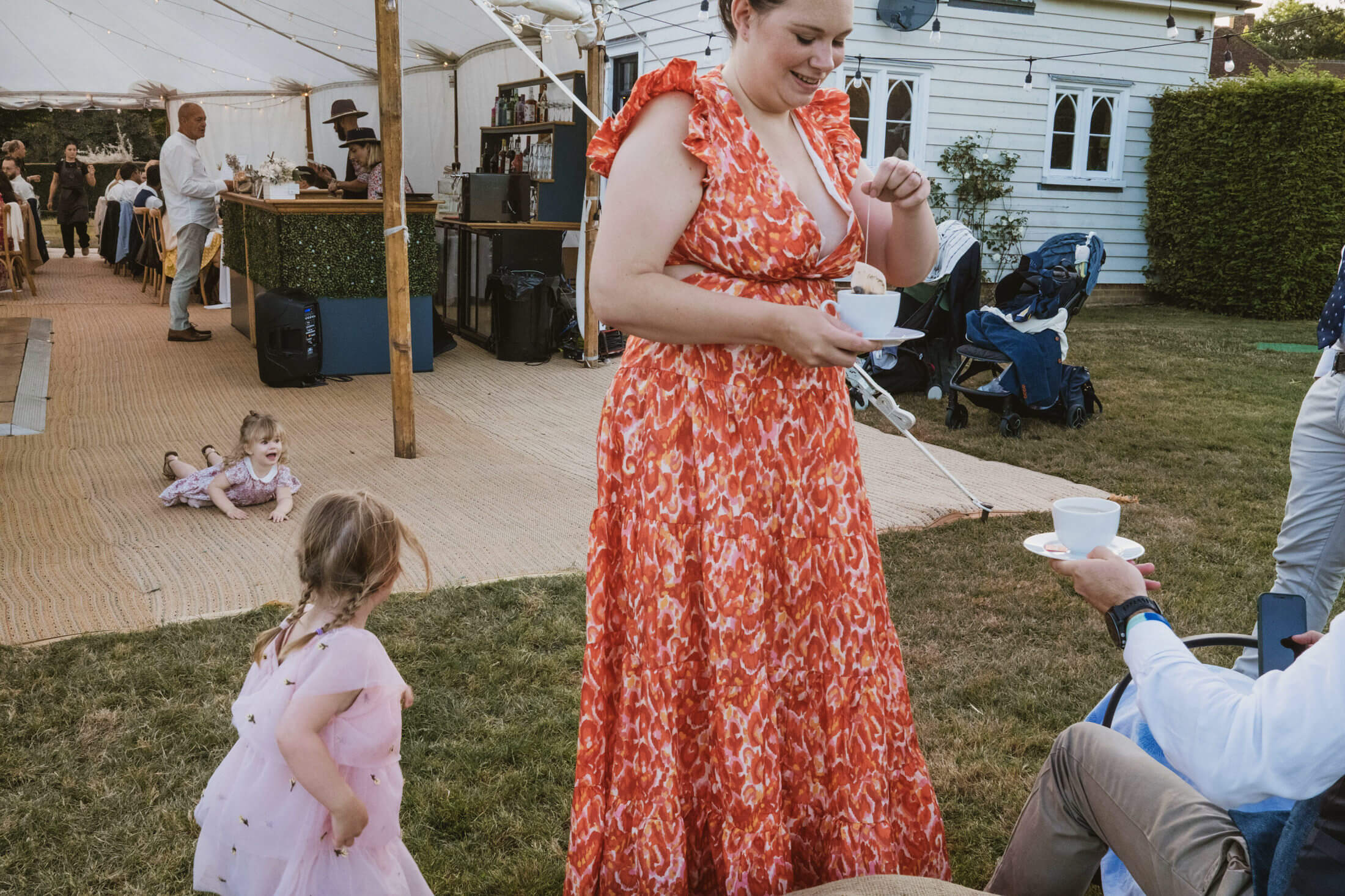 Woman in red dress laughs with tea as children play on lawn during documentary wedding photography.