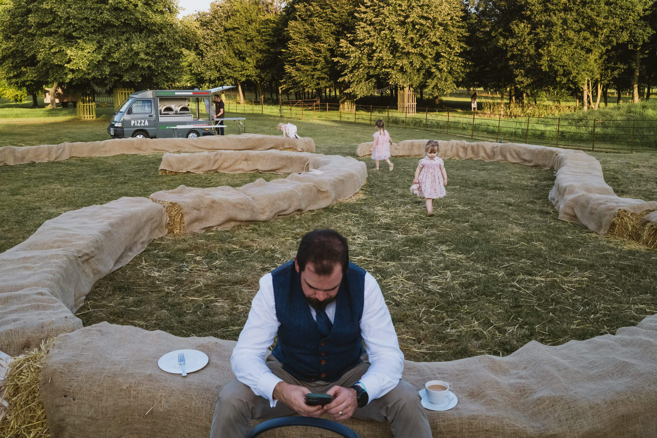 Man in waistcoat checks phone on hay bale as children play near pizza van at wedding reception.
