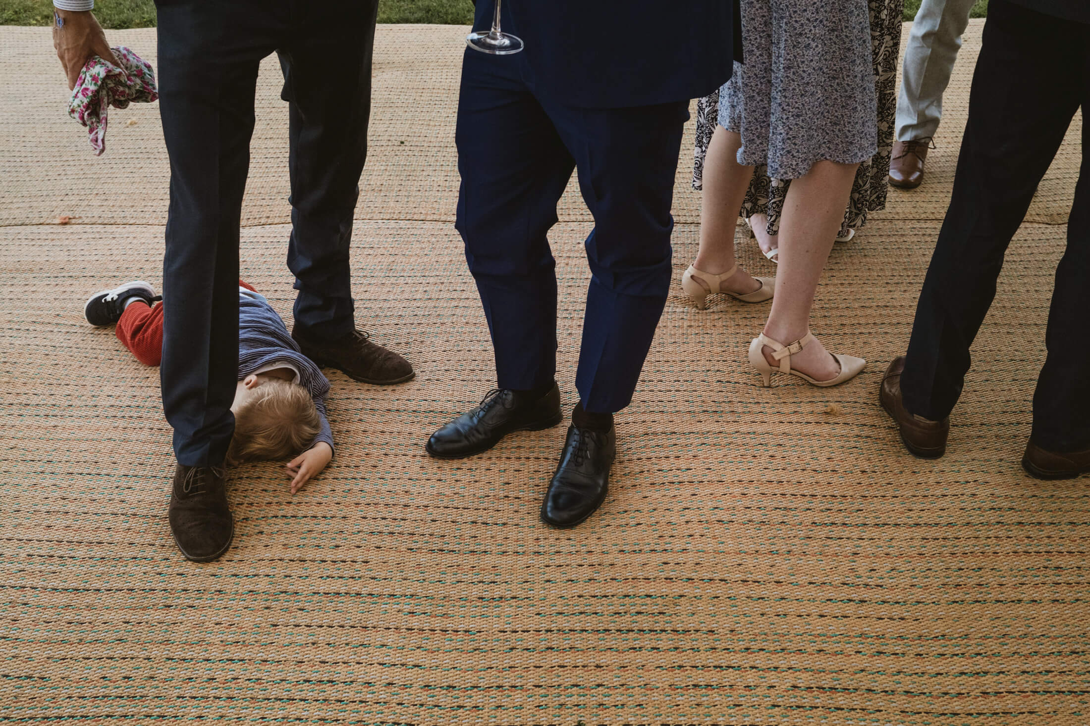 Child lying on woven floor among guests’ legs during wedding reception, a quiet documentary wedding photography moment.