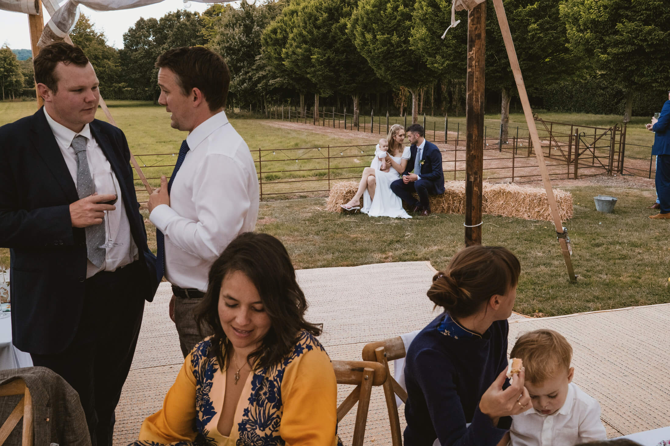 Guests chat beneath a marquee while couple cradle baby on hay bales at countryside wedding reception.