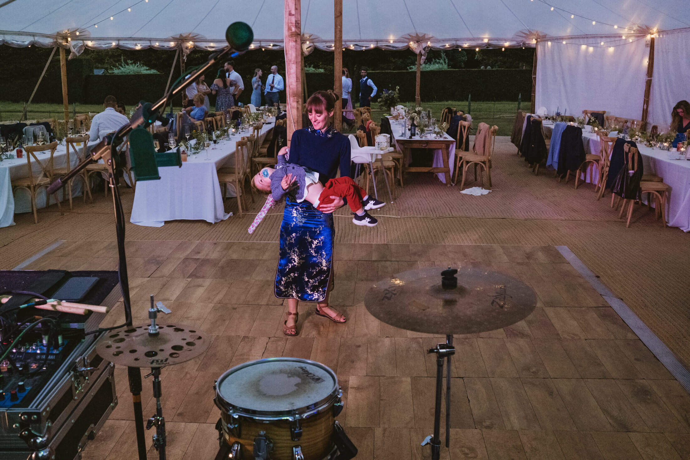 Woman cradles sleeping child on marquee dance floor beside drum kit at wedding reception.