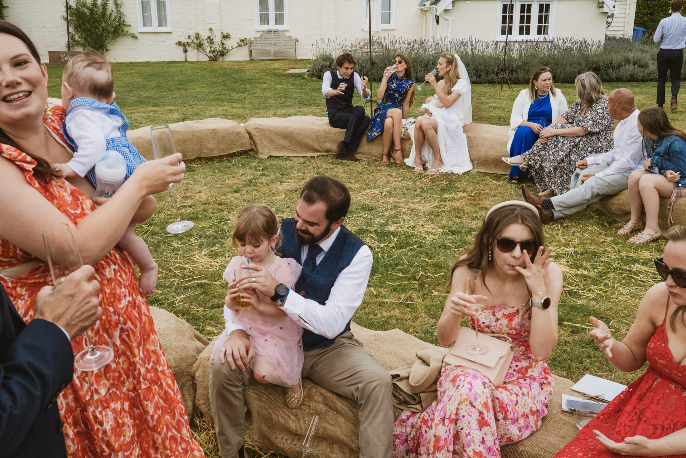 Wedding guests relax on hay bales, chatting and sipping drinks on a sunny lawn, documentary wedding photography.