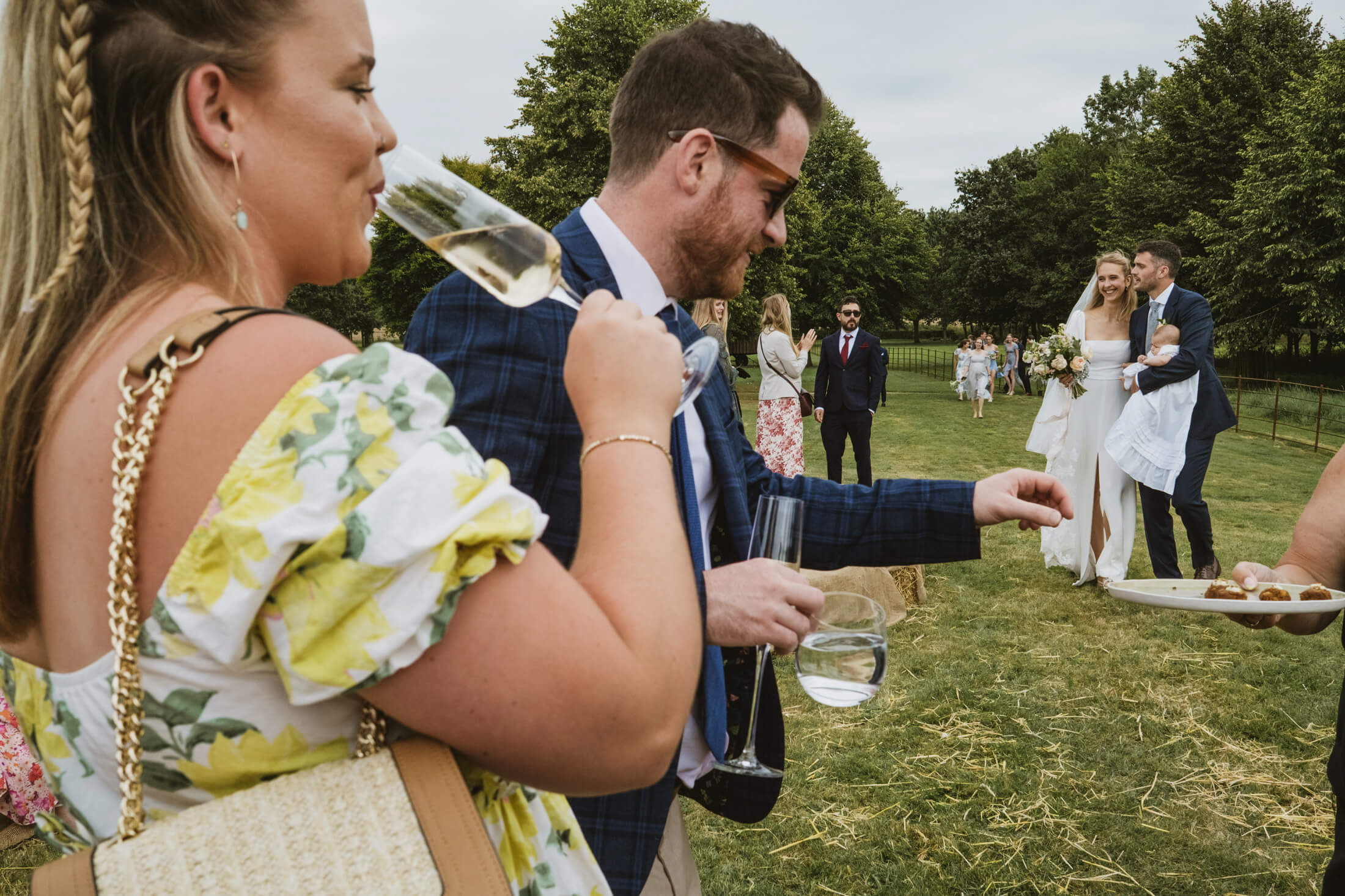 Guests sip champagne as bride and groom walk across lawn with their baby, laughter drifting through the wedding reception.