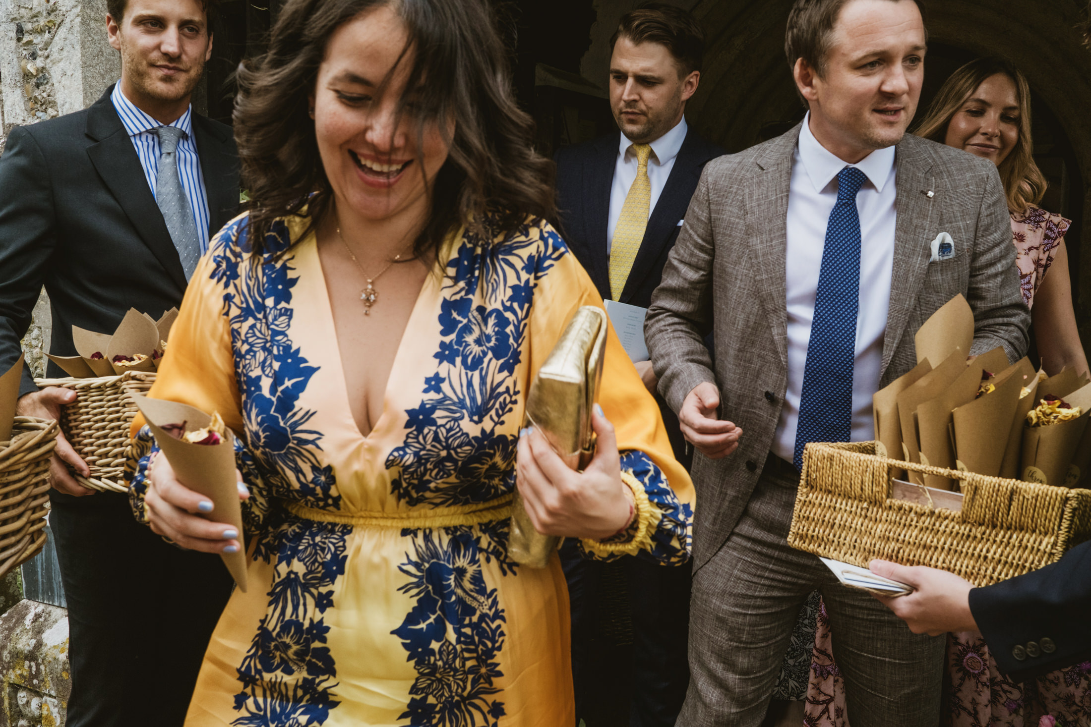 Guest in yellow floral dress laughing, holding confetti cones outside church during documentary wedding photography.