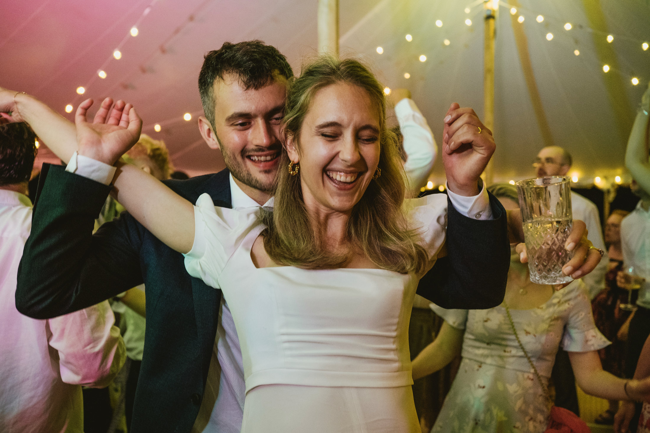 Bride laughing with arms raised as groom embraces her on crowded wedding reception dance floor.