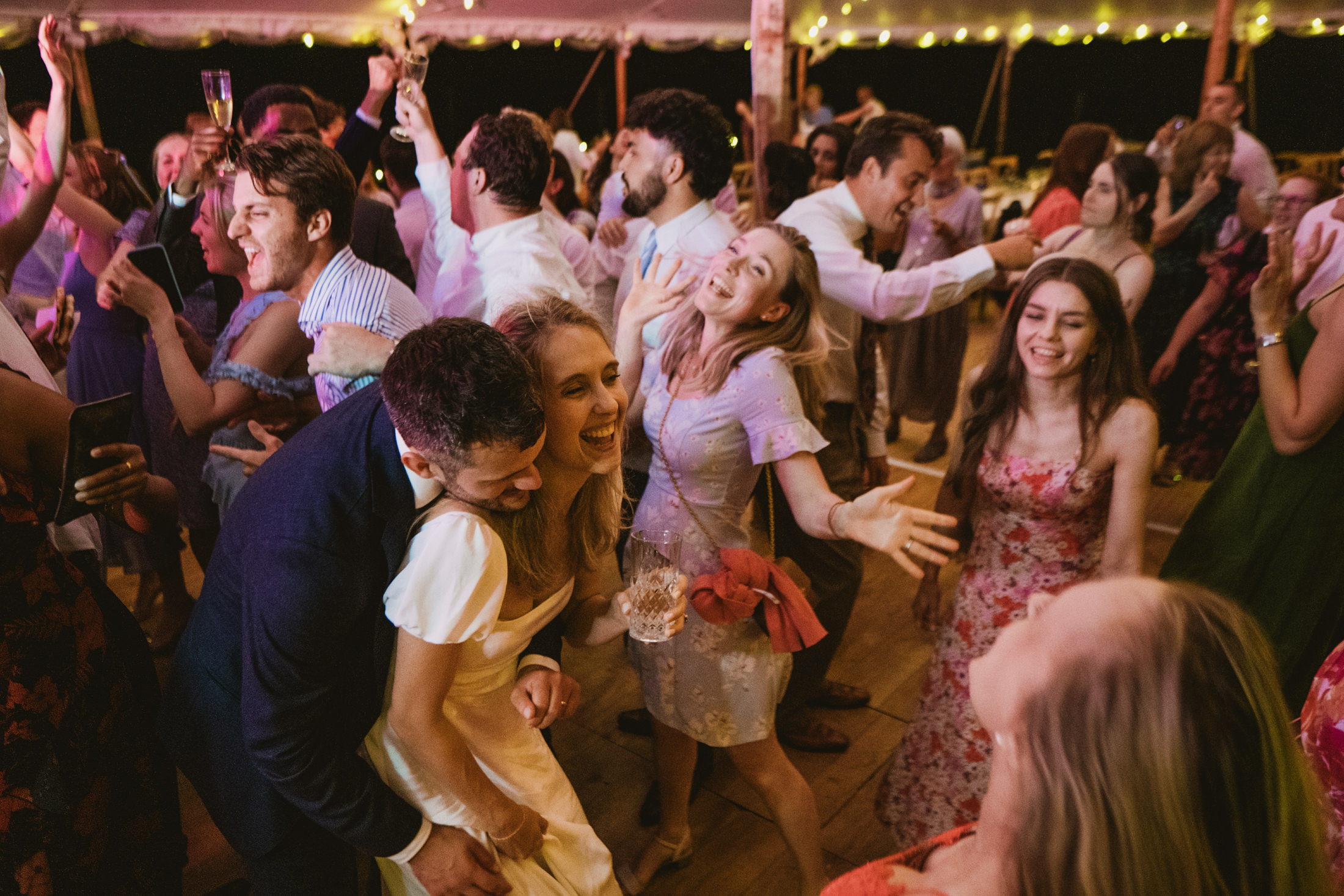 Guests laughing and dancing beneath festoon lights at a lively wedding reception.