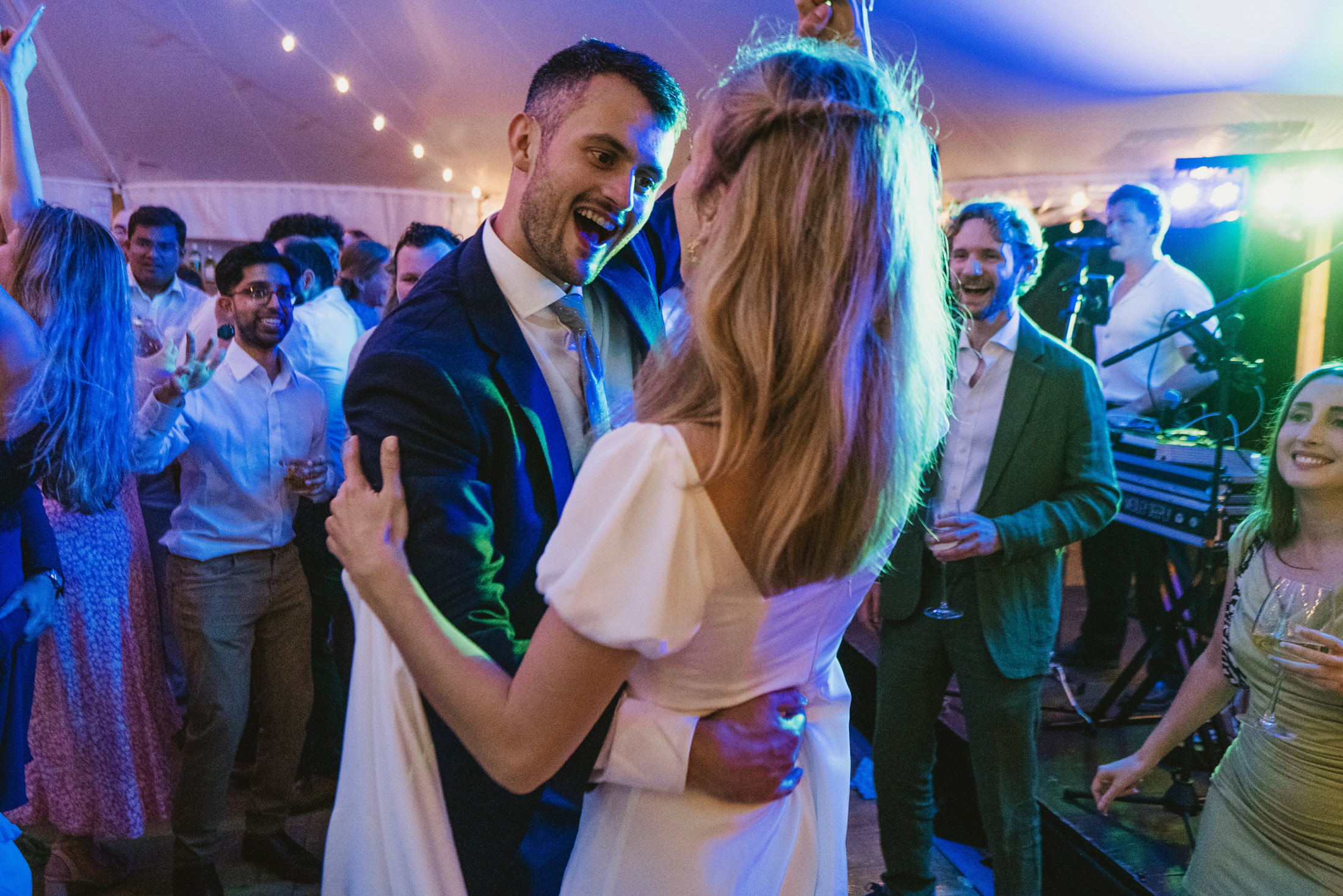 Bride and groom laughing as they dance among friends at a lively wedding reception.