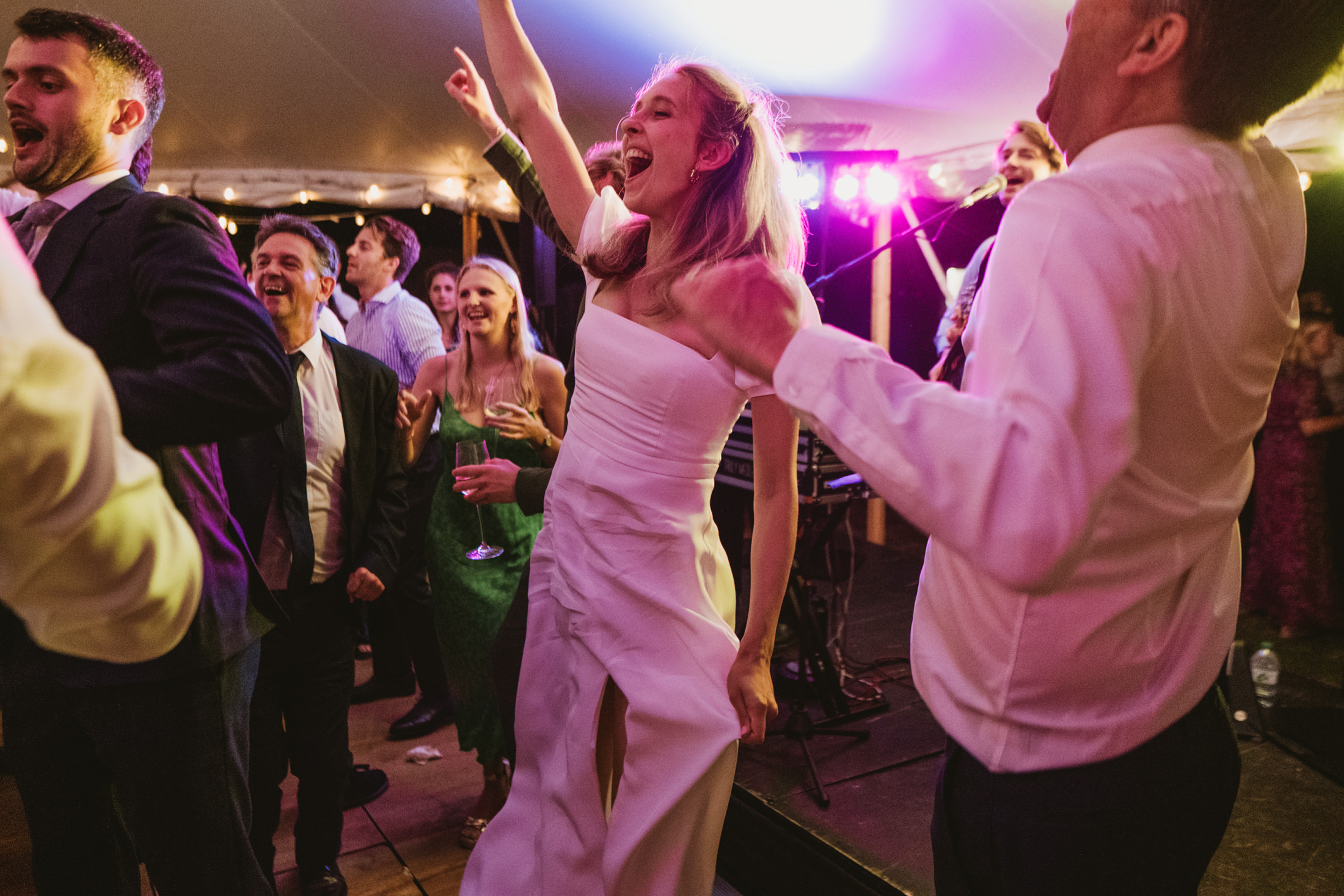Bride laughing with arm raised as guests dance beneath glowing lights at a wedding reception.