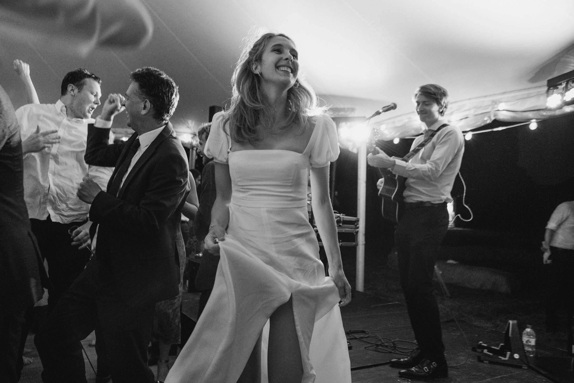Bride laughing as she dances, guests cheering beside live band during evening wedding reception.