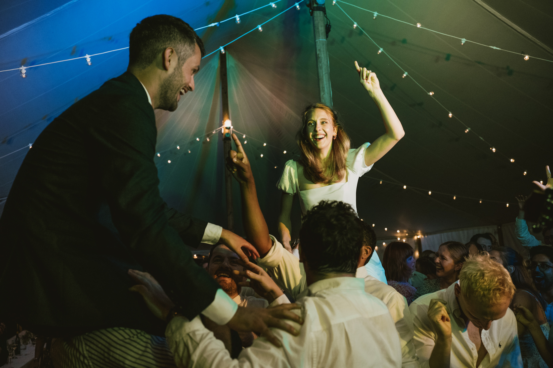 Bride and groom lifted above cheering guests on a crowded wedding reception dance floor beneath fairy lights.