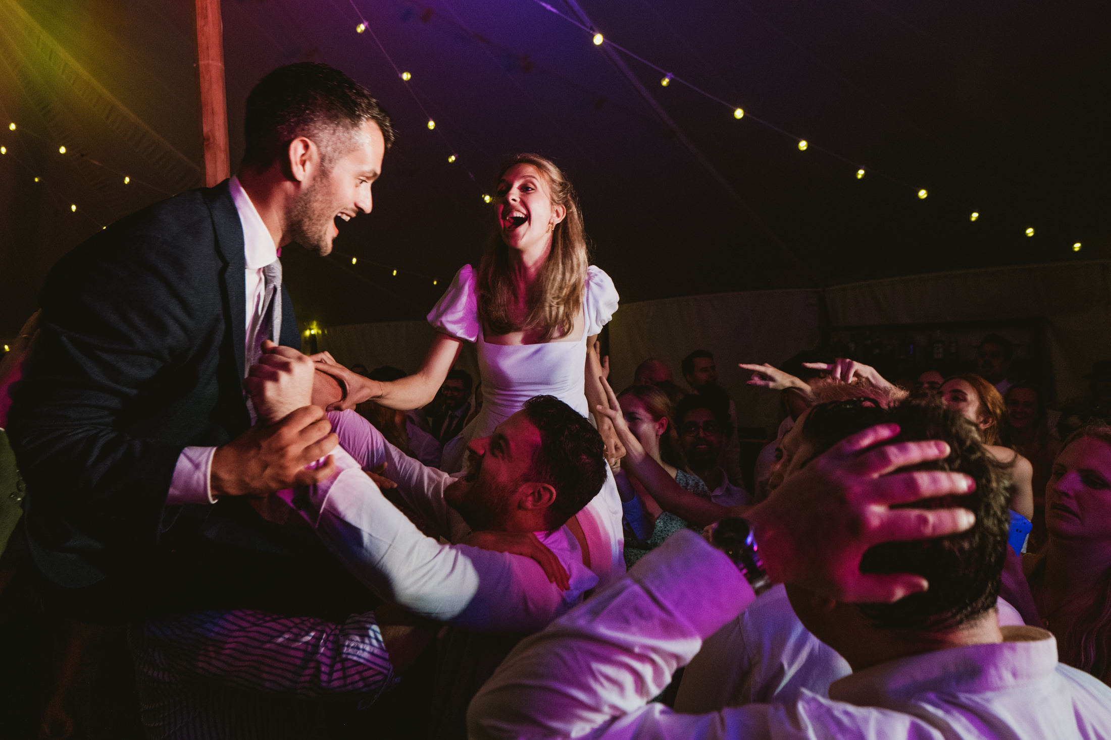 Bride and groom lifted above cheering guests, laughing beneath fairy lights during lively wedding reception.