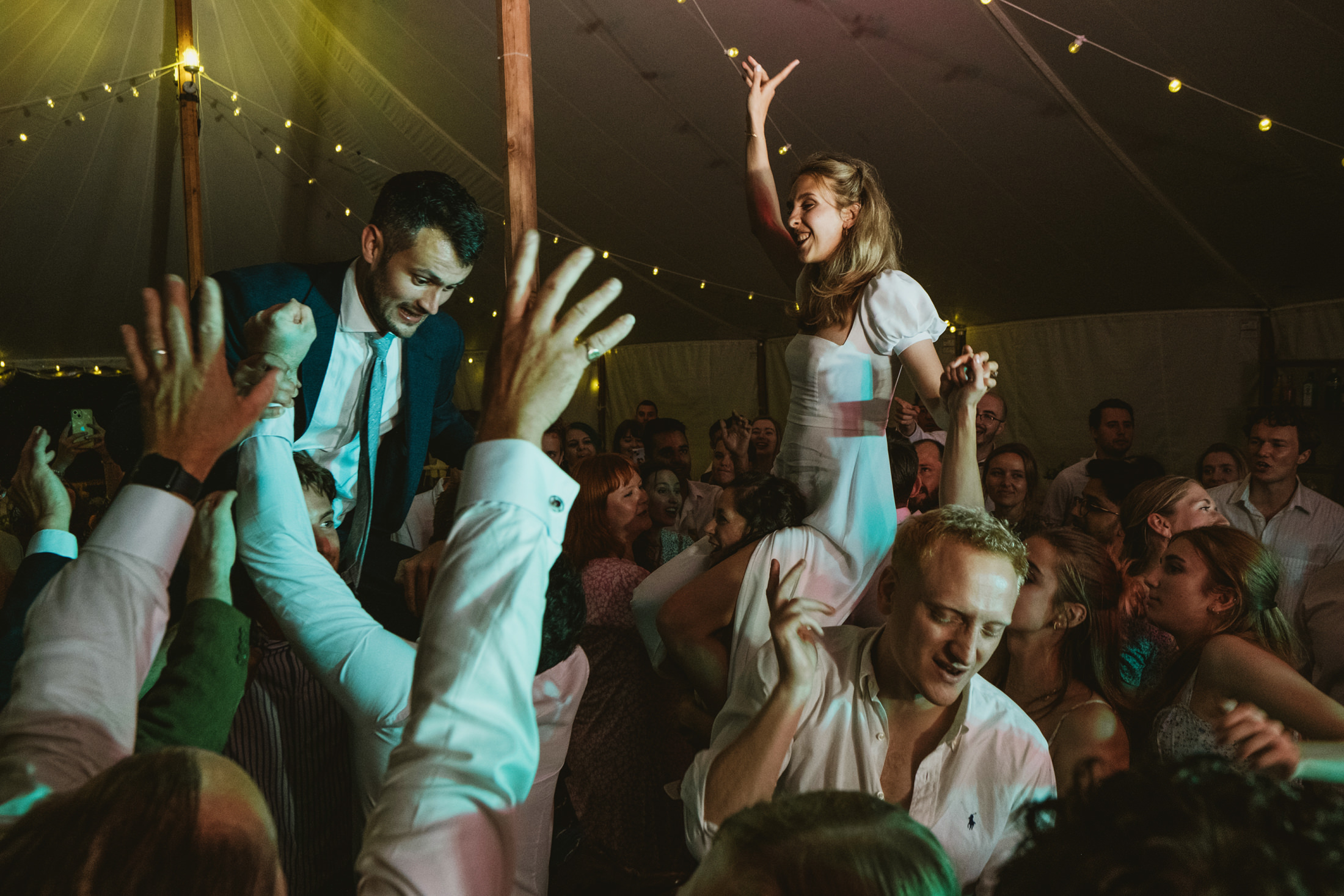 Bride and groom lifted above cheering guests on a crowded dance floor beneath warm festoon lights.
