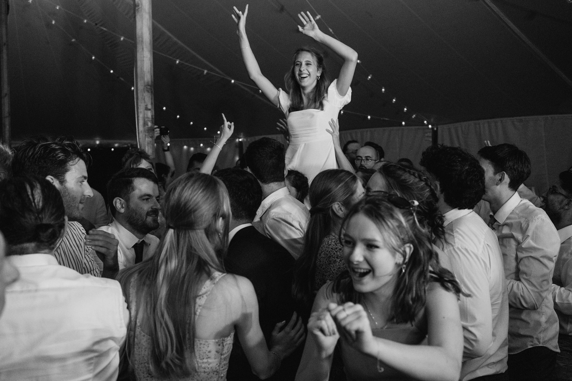 Bride lifted above dancing guests at wedding reception, arms raised beneath glowing marquee lights.