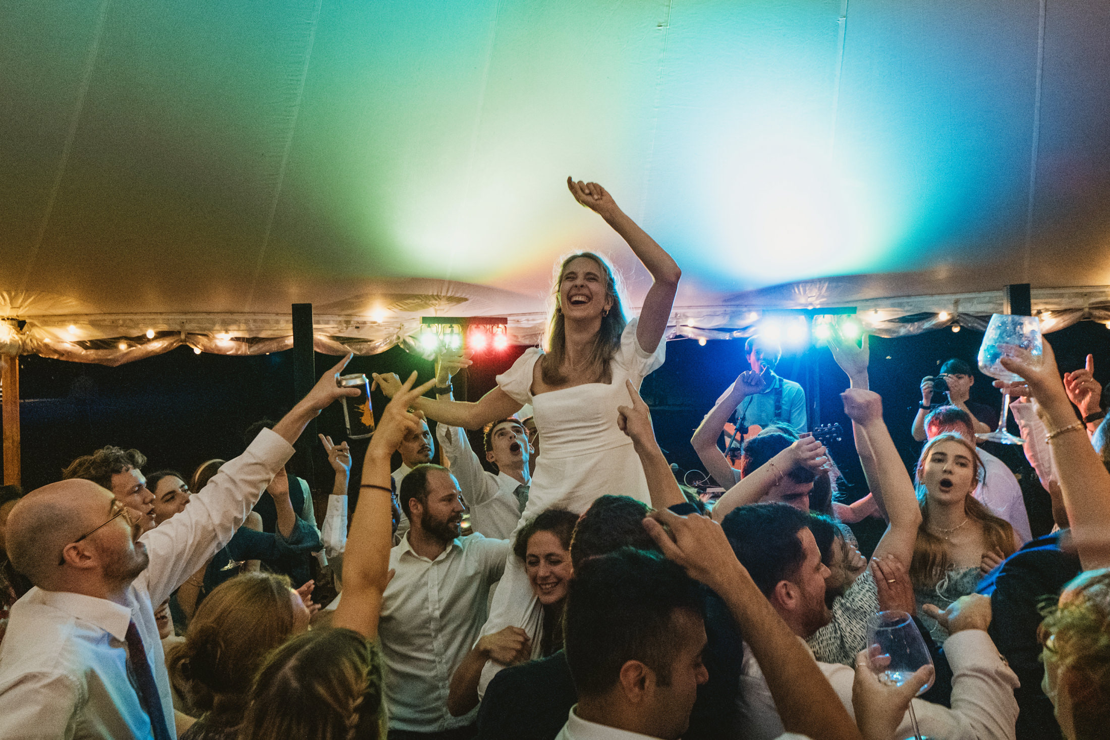 Bride lifted above cheering guests on a crowded dance floor beneath colourful wedding reception lights.