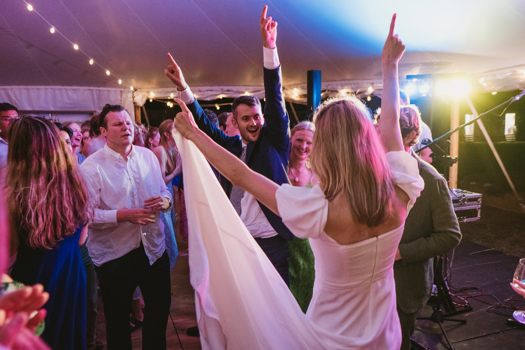 Bride and groom with raised arms dancing among guests beneath festoon lights, documentary wedding photography.