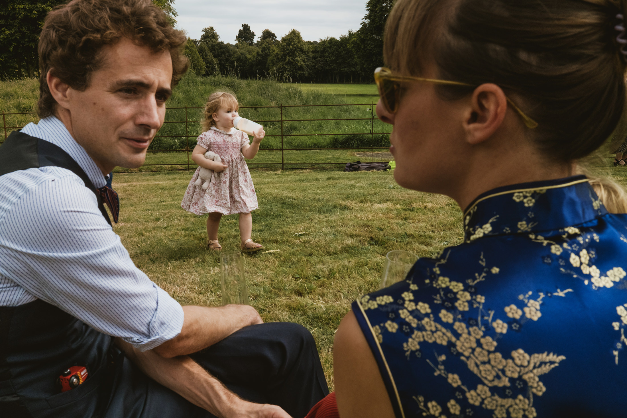 Parents watch toddler sipping from bottle and clutching teddy on sunlit lawn, documentary wedding photography.