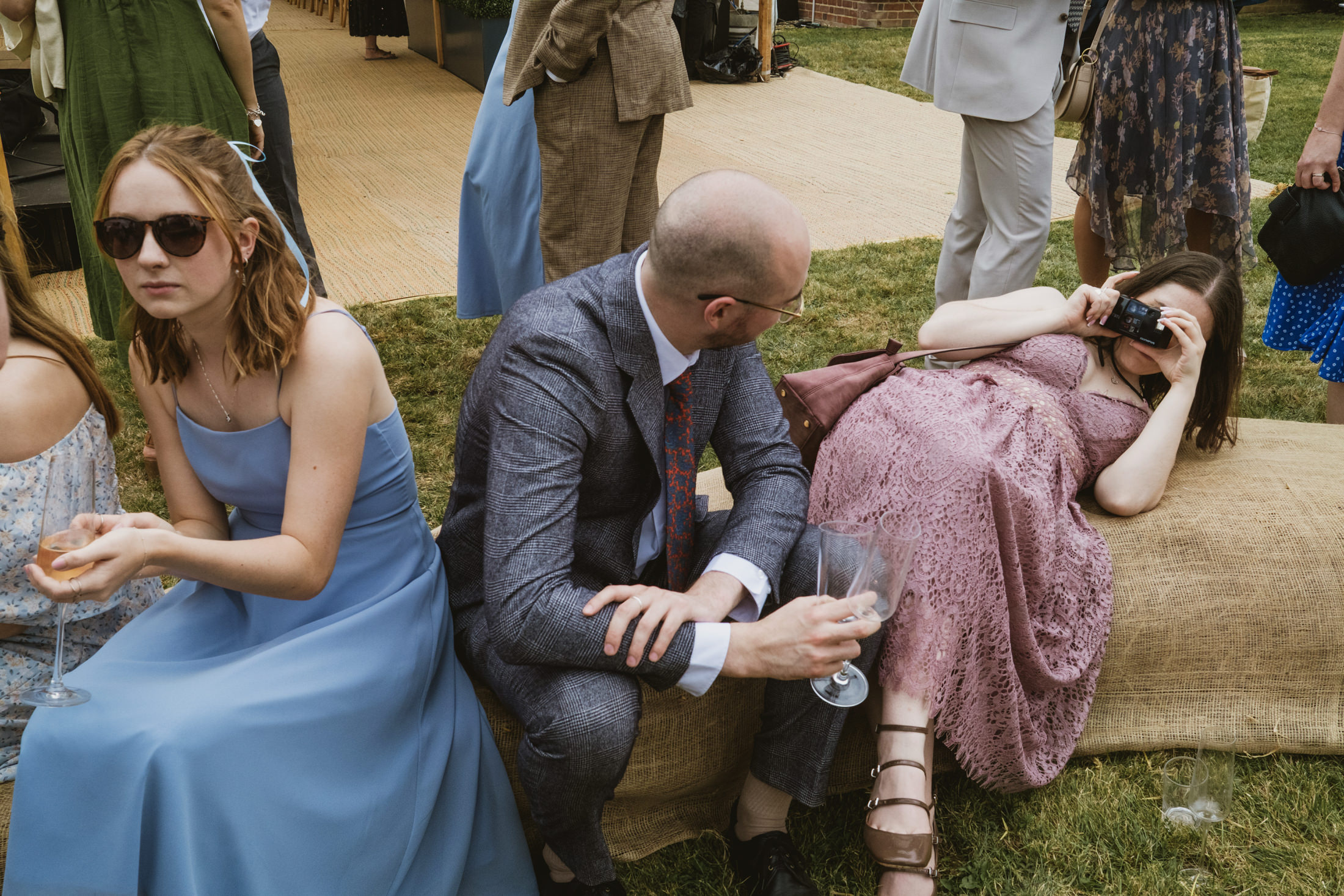 Guests relax on grass during outdoor wedding, woman lying back taking photo, friends holding drinks in documentary wedding photography.