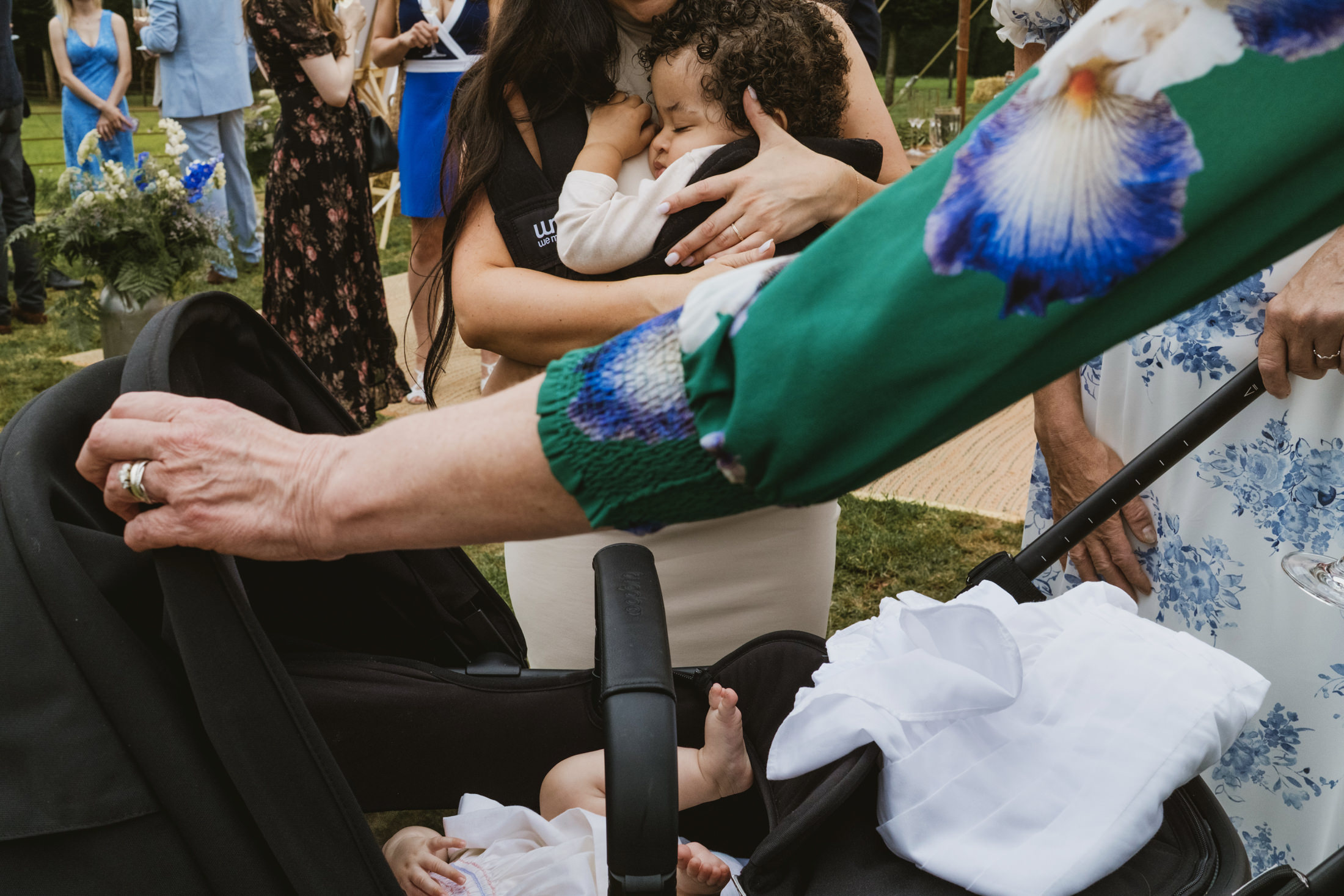 Woman cradles sleeping toddler beside pram at outdoor wedding reception.