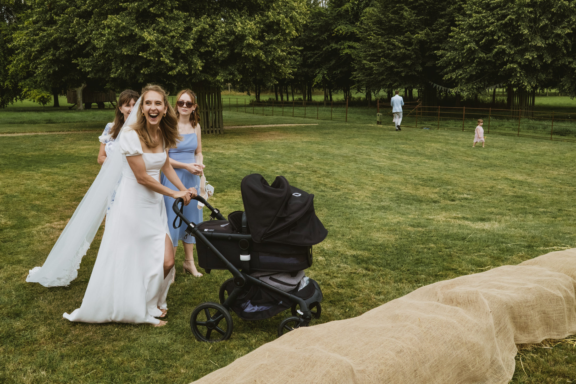 Bride laughing while pushing pram across lawn with bridesmaids, candid wedding photography moment.