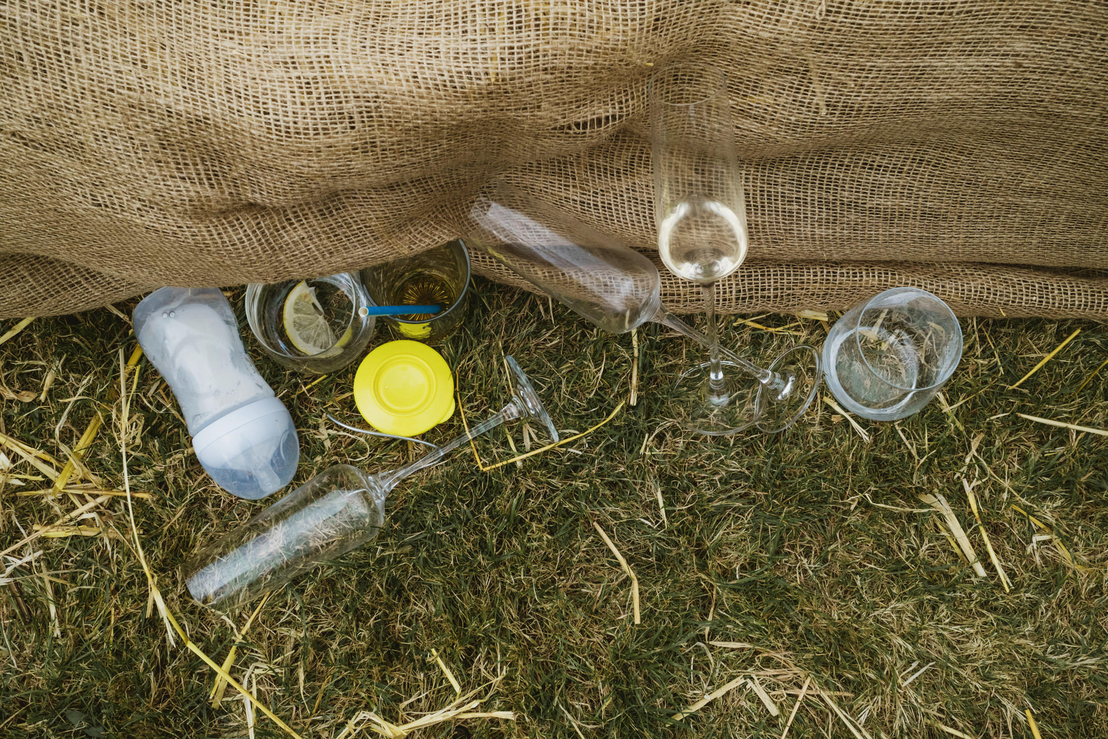 Empty champagne flutes and bottles on grass beneath burlap at outdoor wedding reception.