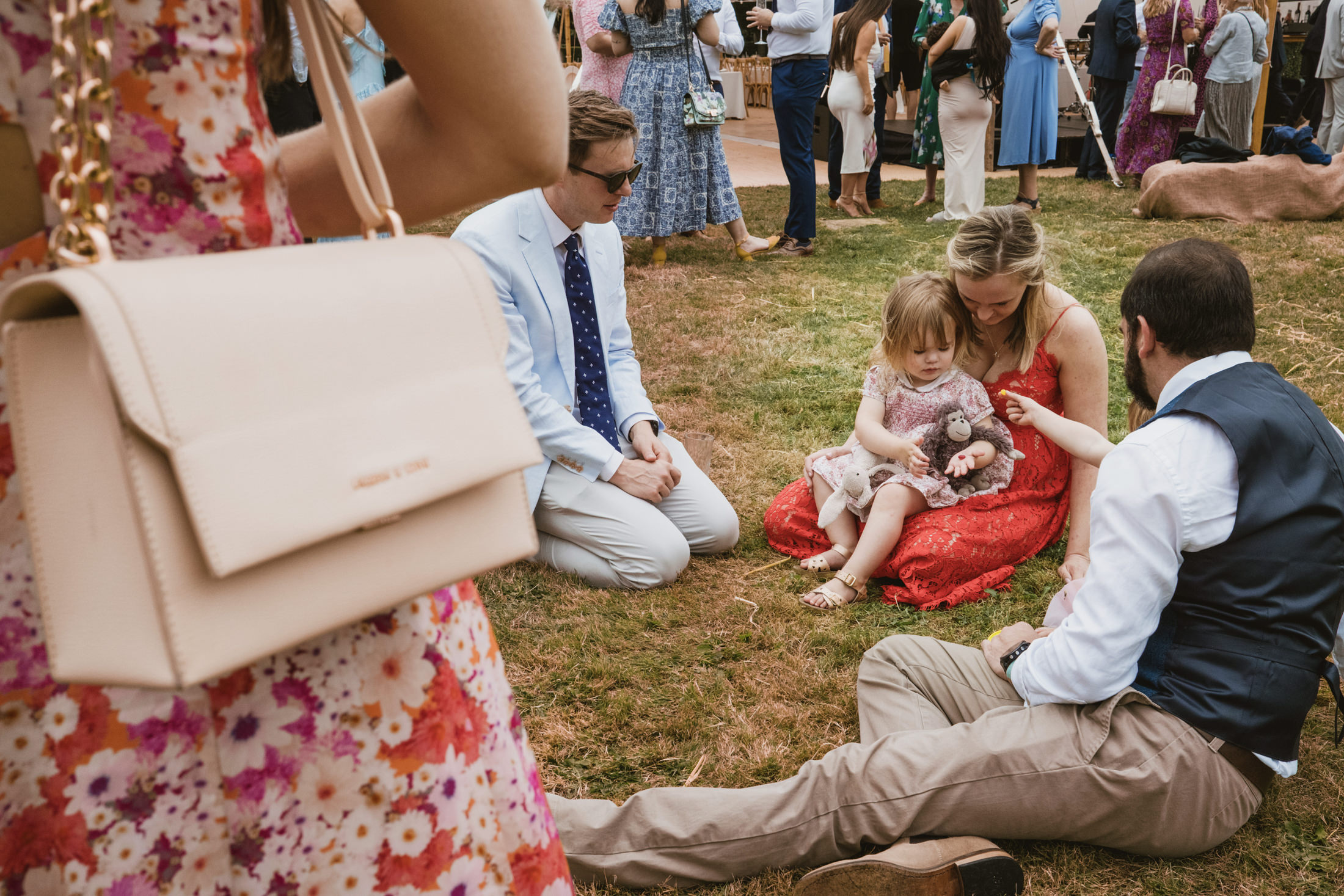 Outdoor wedding reception scene in documentary wedding photography style, guests sit on grass with toddler and toy.