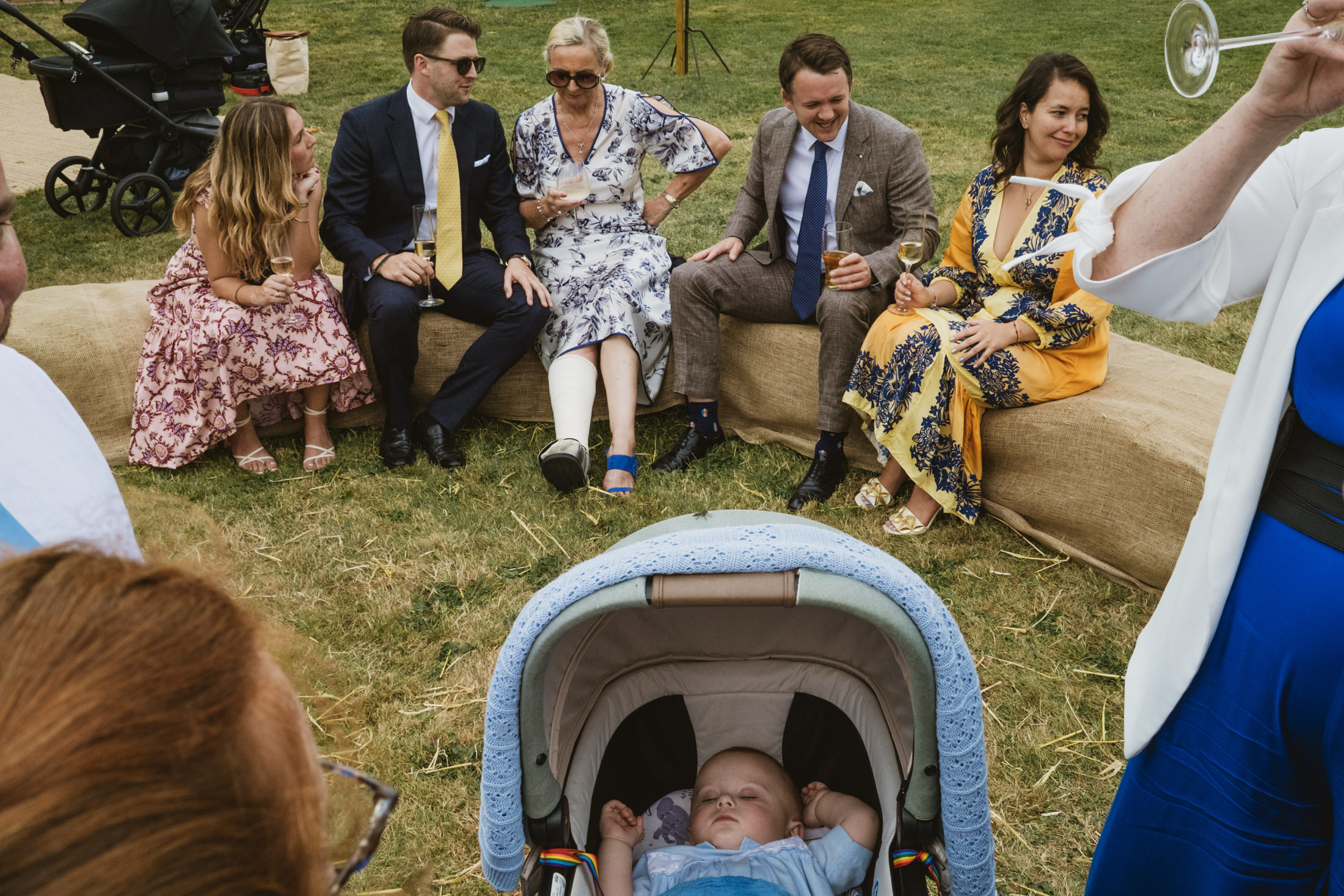 Wedding guests laughing on a garden bench as a baby sleeps in a pram nearby.