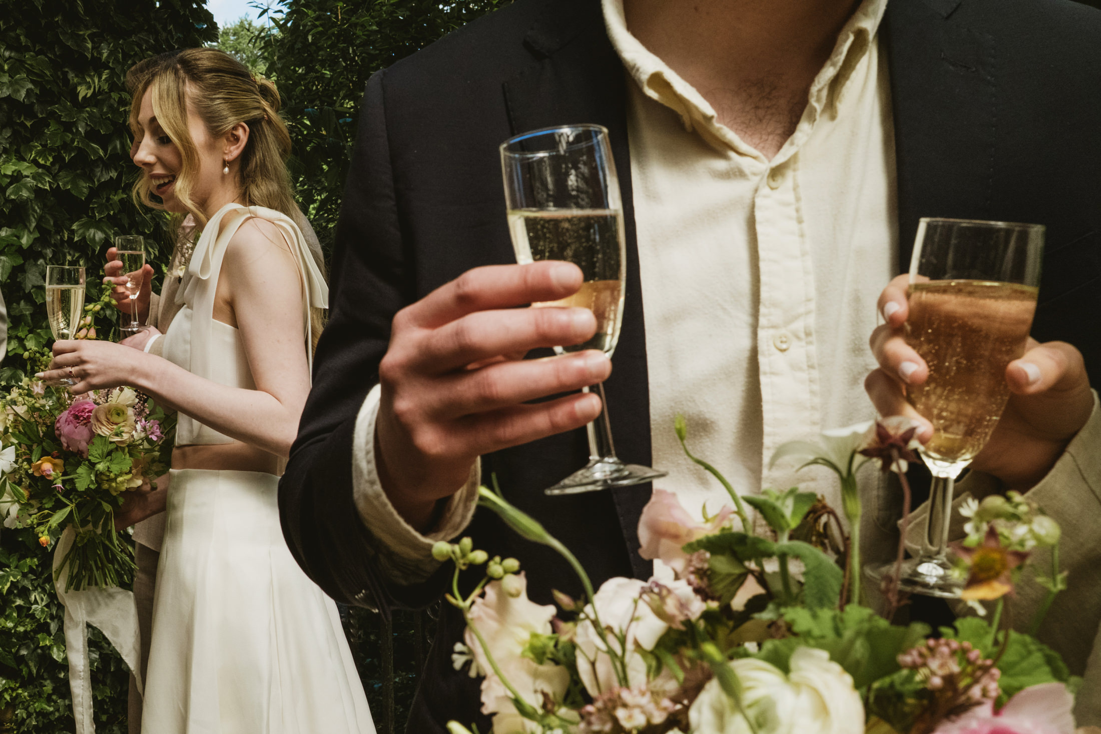 Guests laugh with champagne flutes beside bride holding bouquet in garden wedding reception.