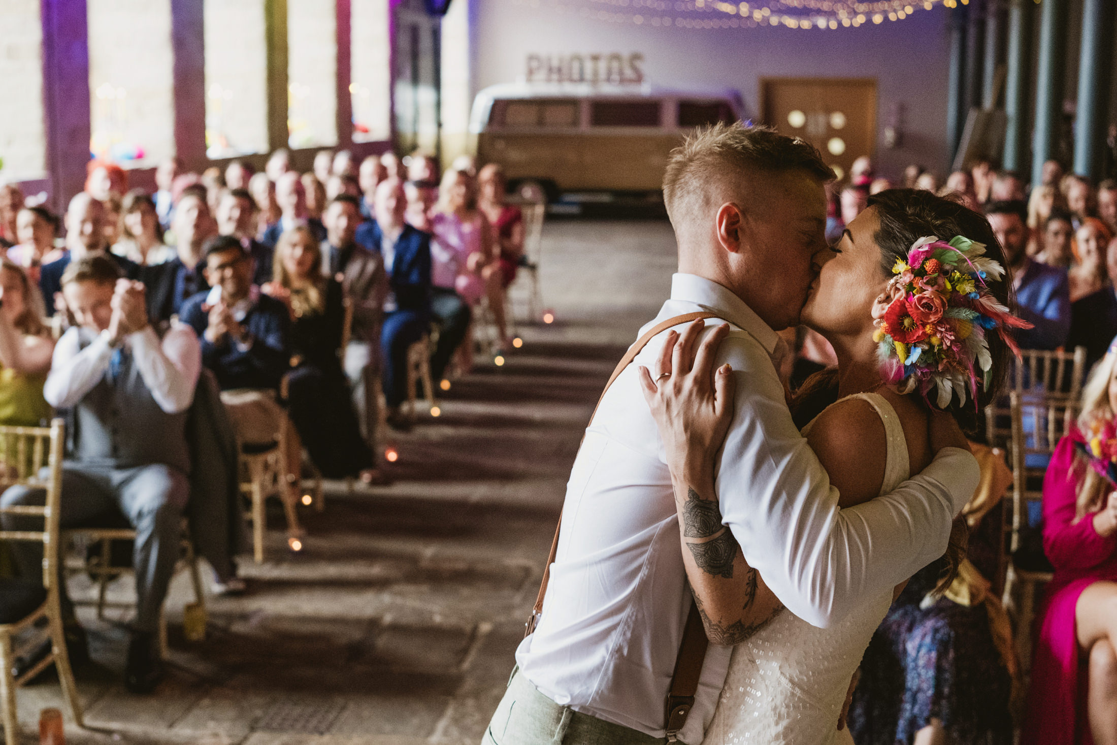 Couple kiss during wedding ceremony as guests applaud in softly lit hall.