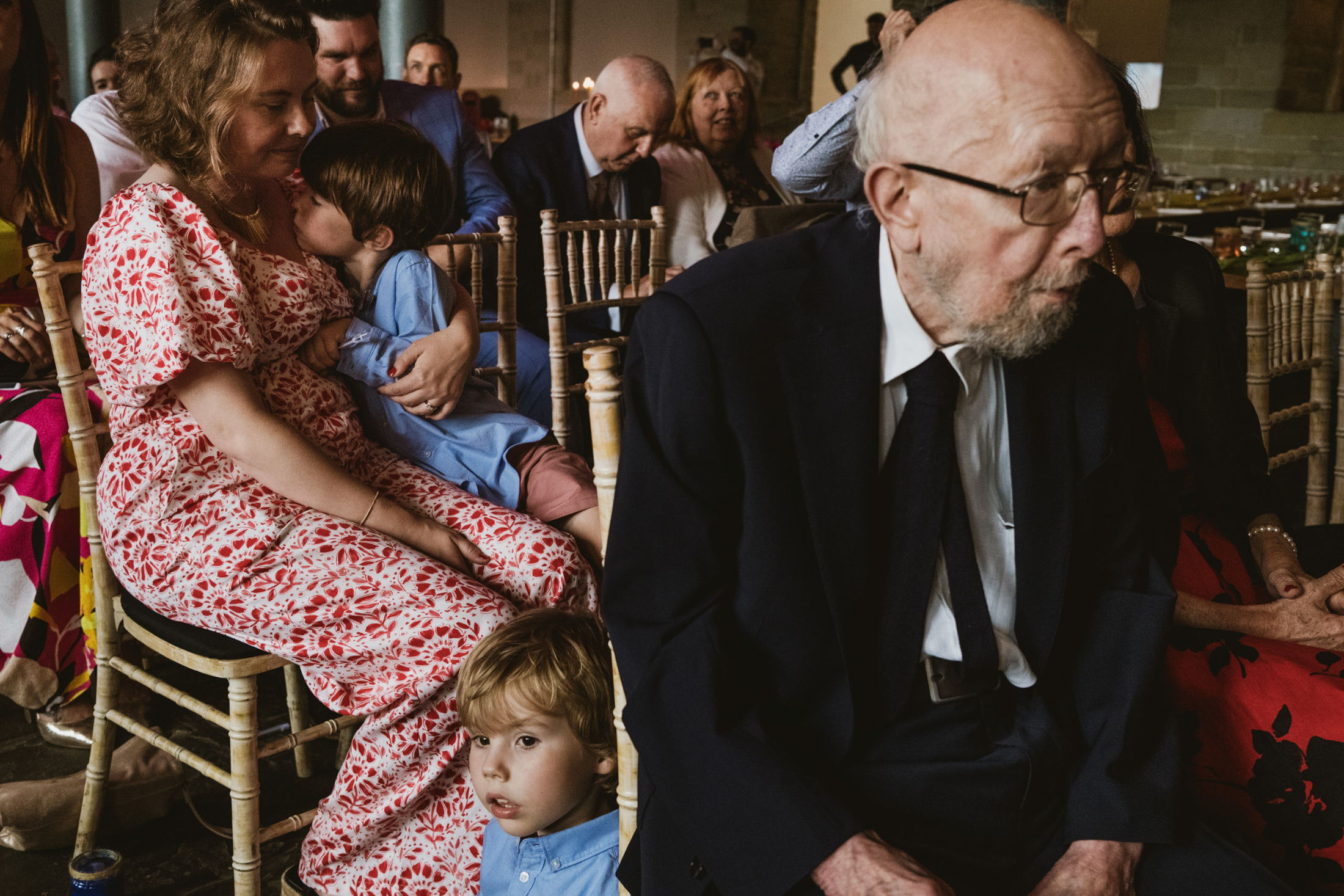 Wedding guests seated indoors, woman cradling child as elderly man looks down, quiet family moment.