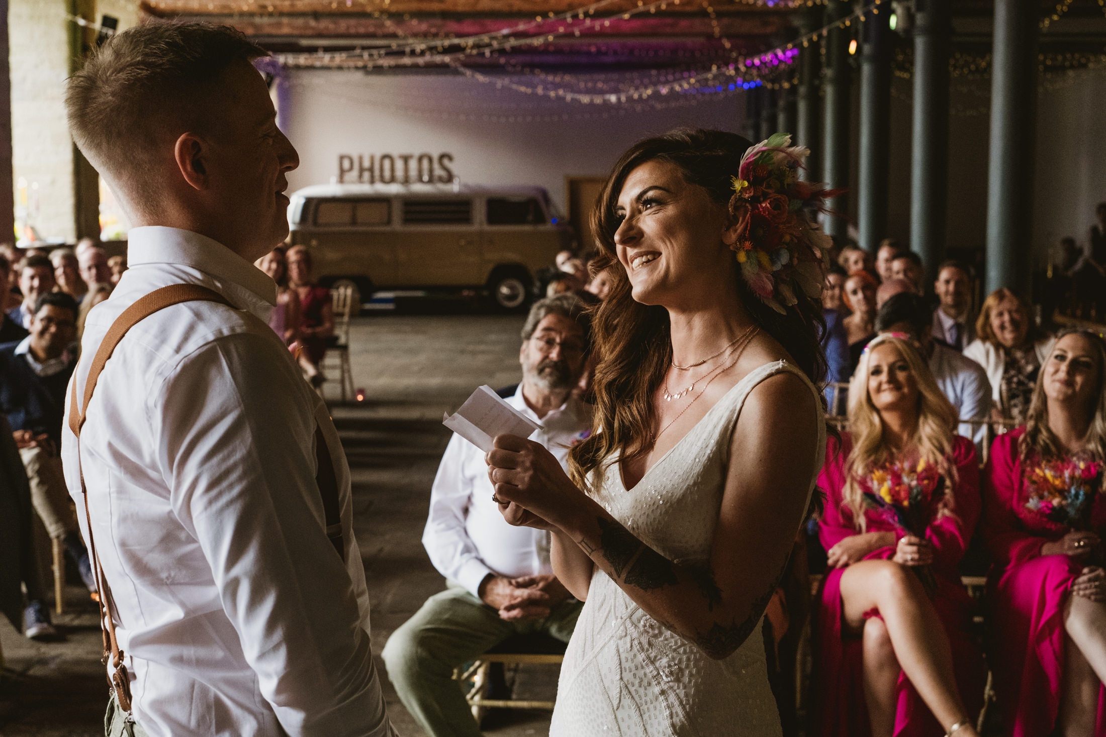 Bride smiles at groom while reading vows, guests watching softly during wedding ceremony.