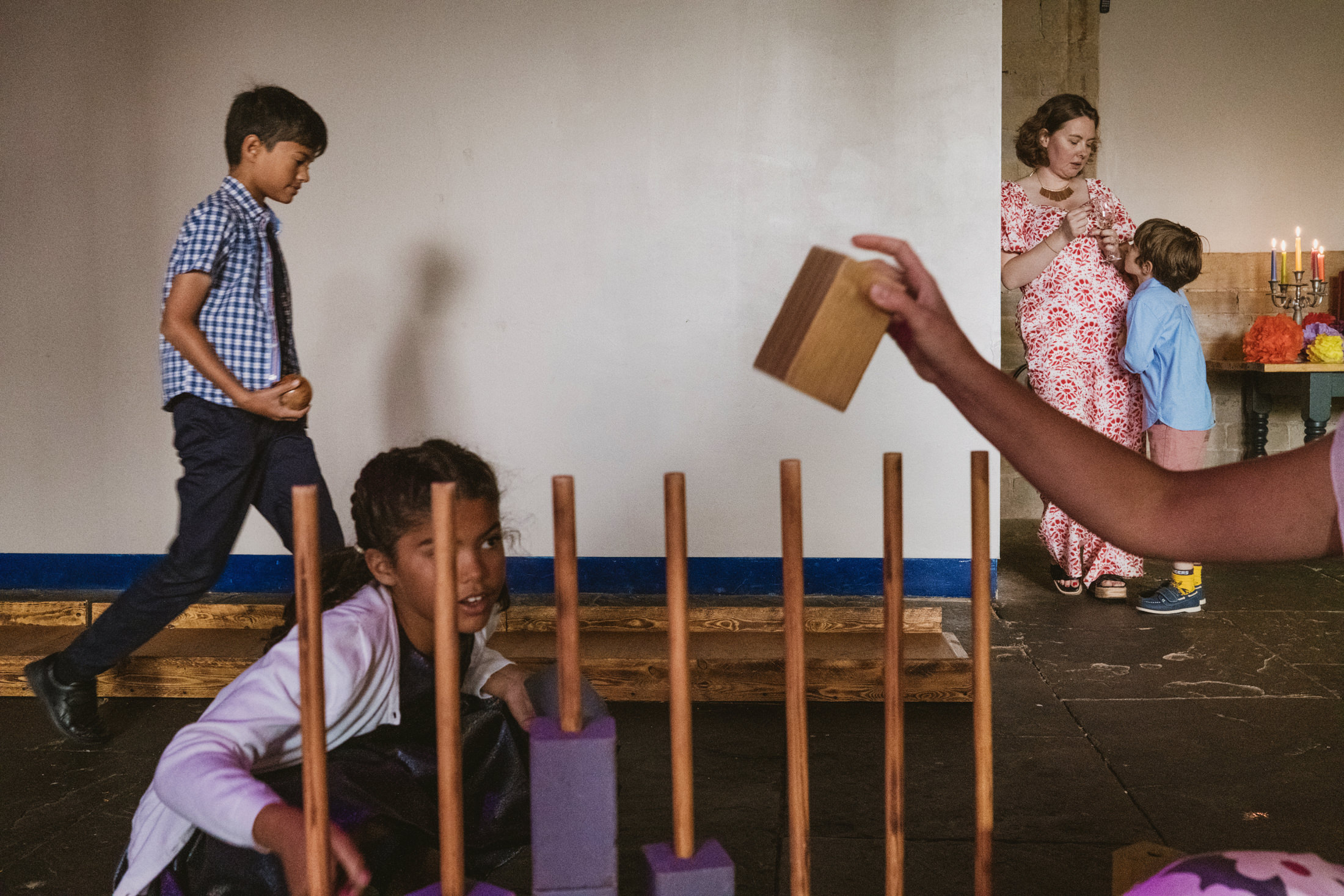 Children play traditional game indoors while woman lights candles in background at wedding celebration.