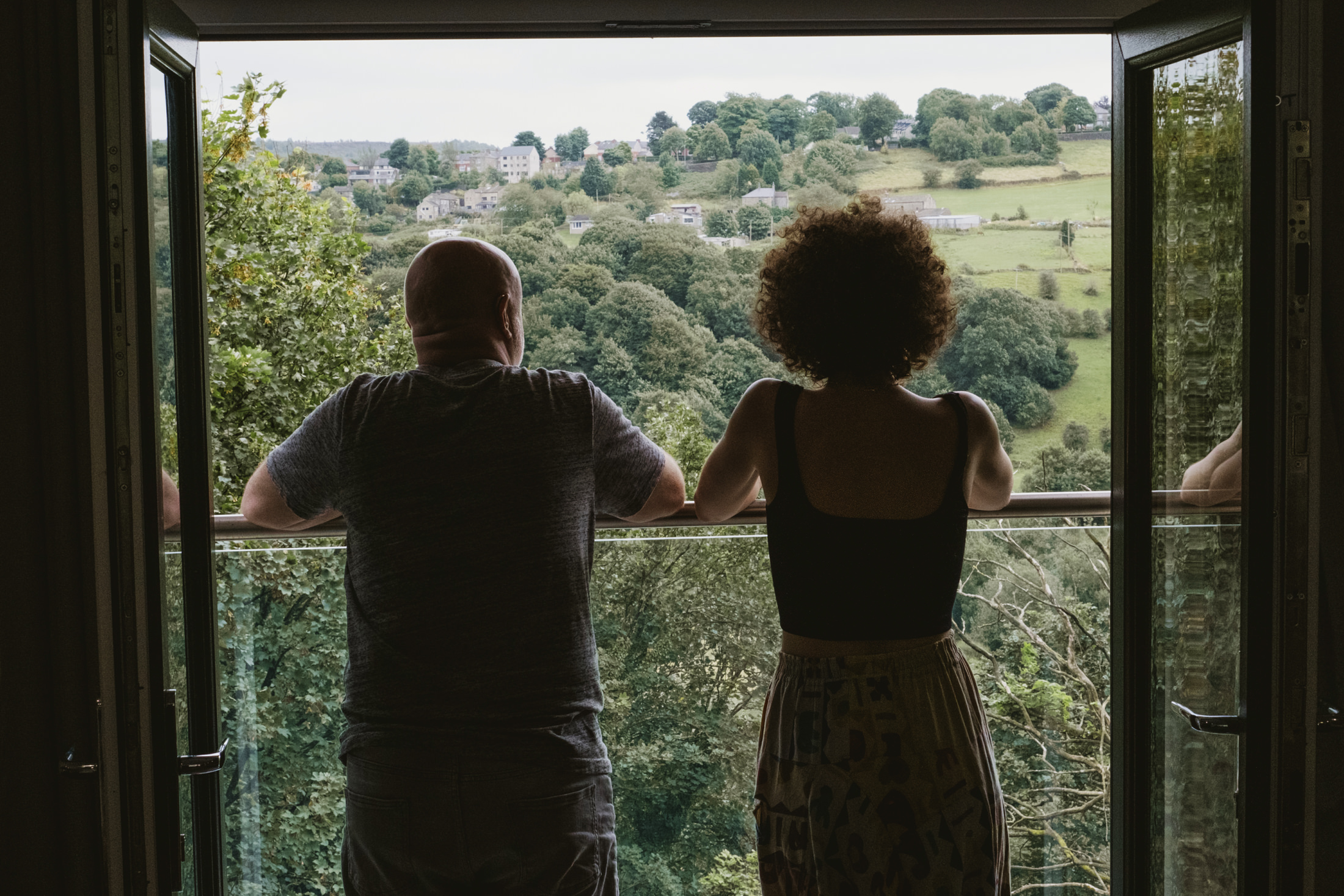 Two people lean on a balcony, gazing over rolling green hills through open doors.