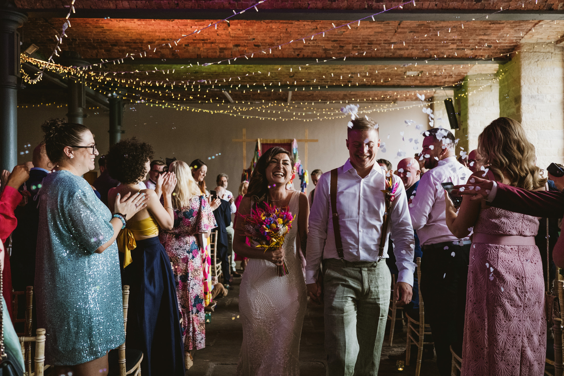 Bride and groom walk through cheering guests as confetti falls during wedding ceremony.