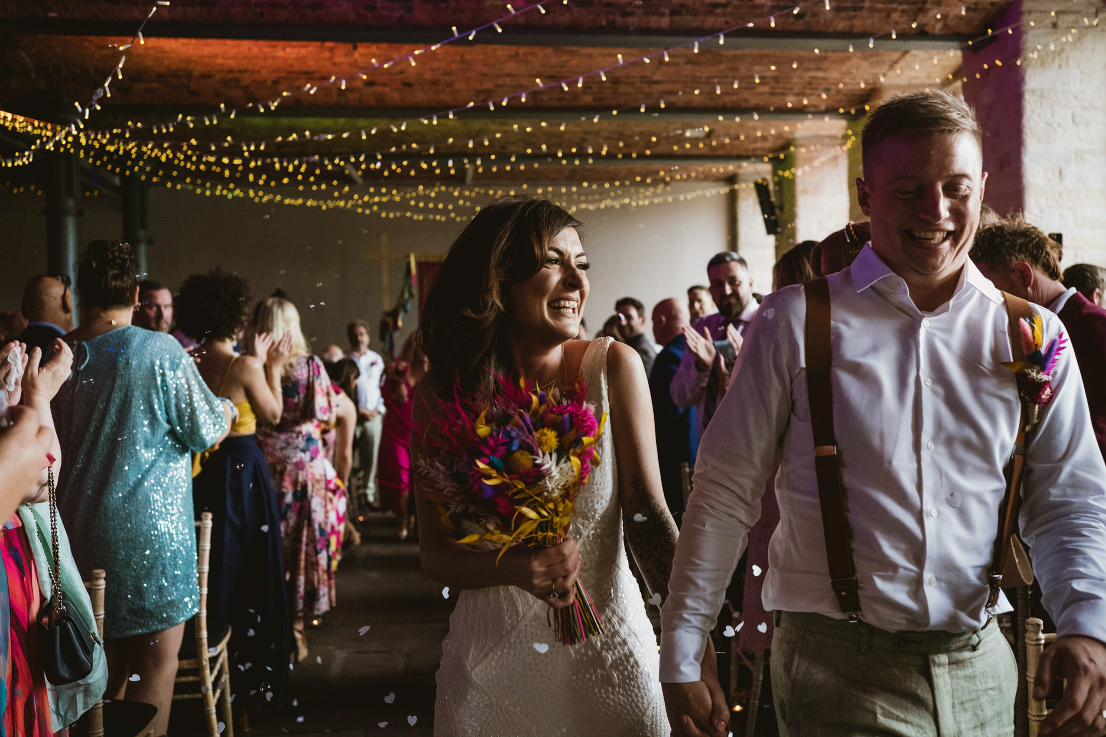 Bride and groom laughing beneath fairy lights as guests toss confetti during barn wedding documentary wedding photography moment.