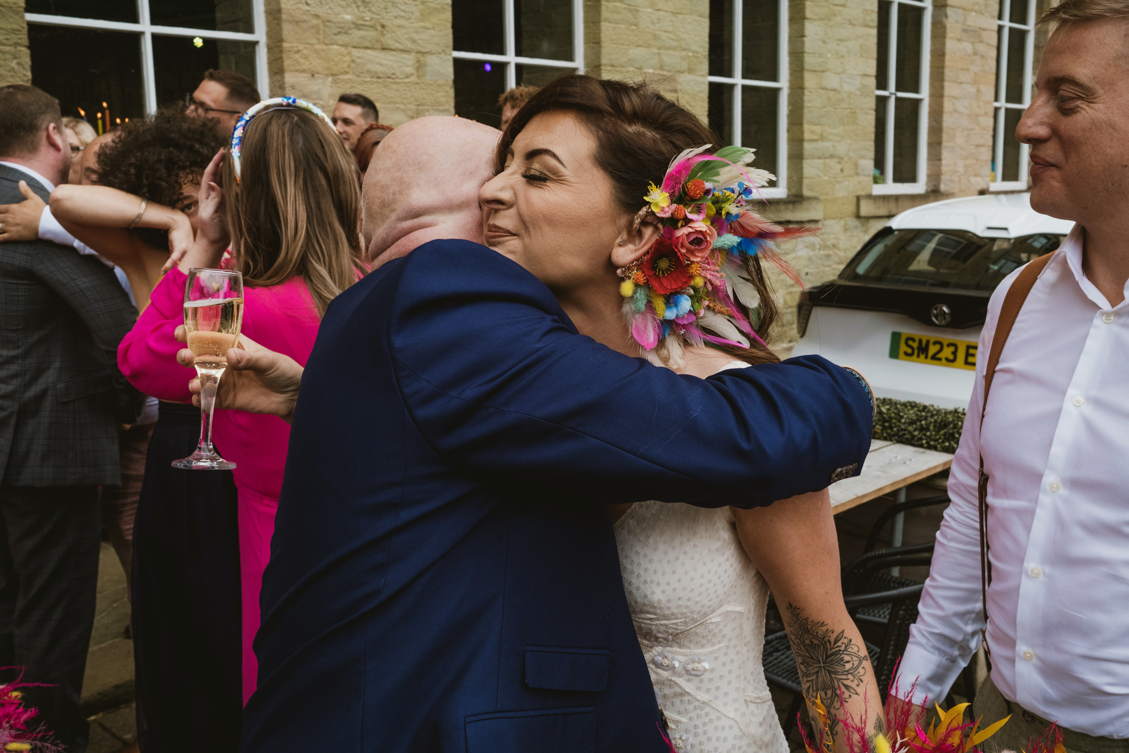 Bride hugs guest holding champagne during joyful wedding reception outside stone venue.