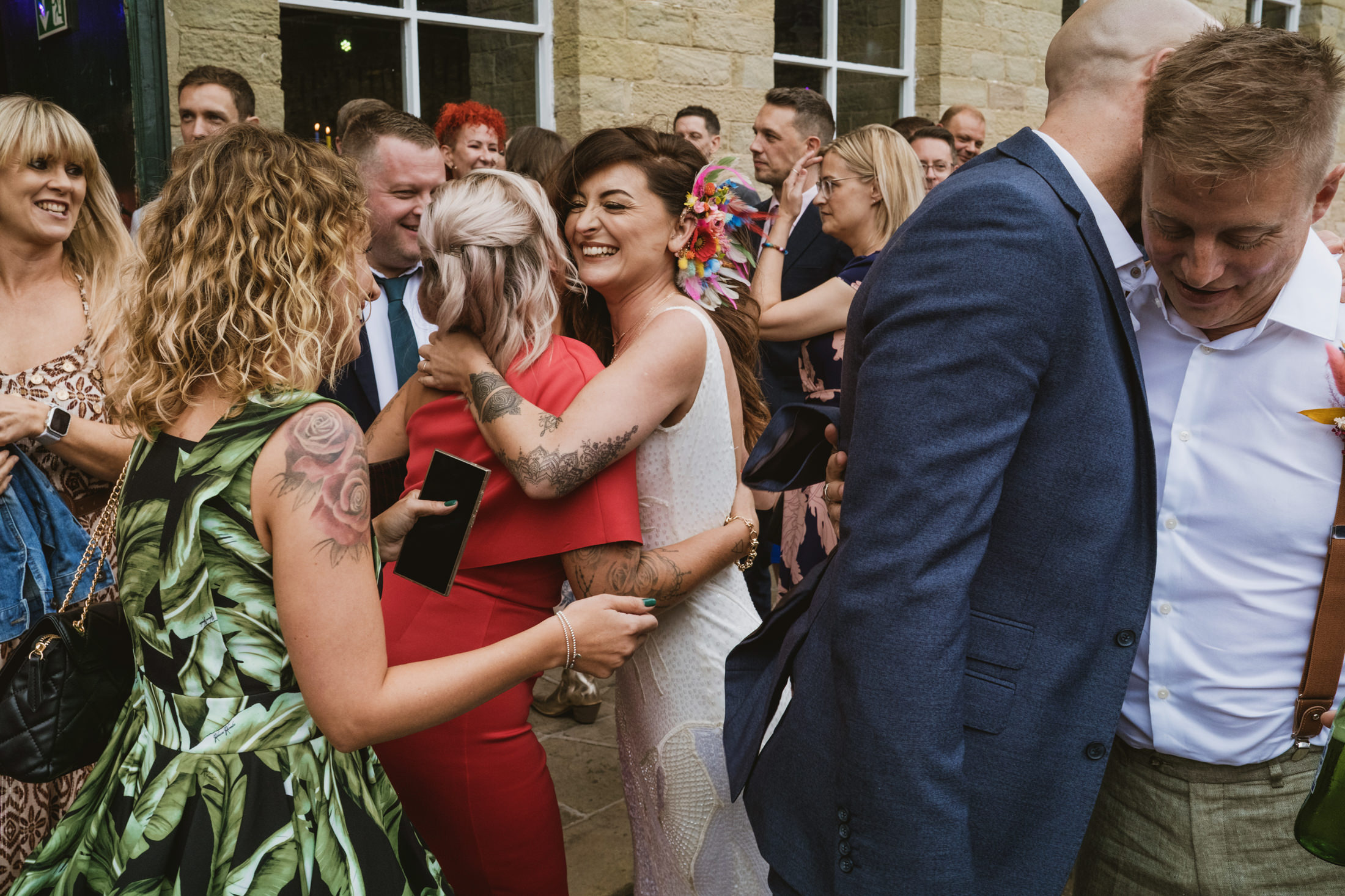 Bride laughing as she hugs a guest outside stone venue, surrounded by smiling friends during wedding photography.