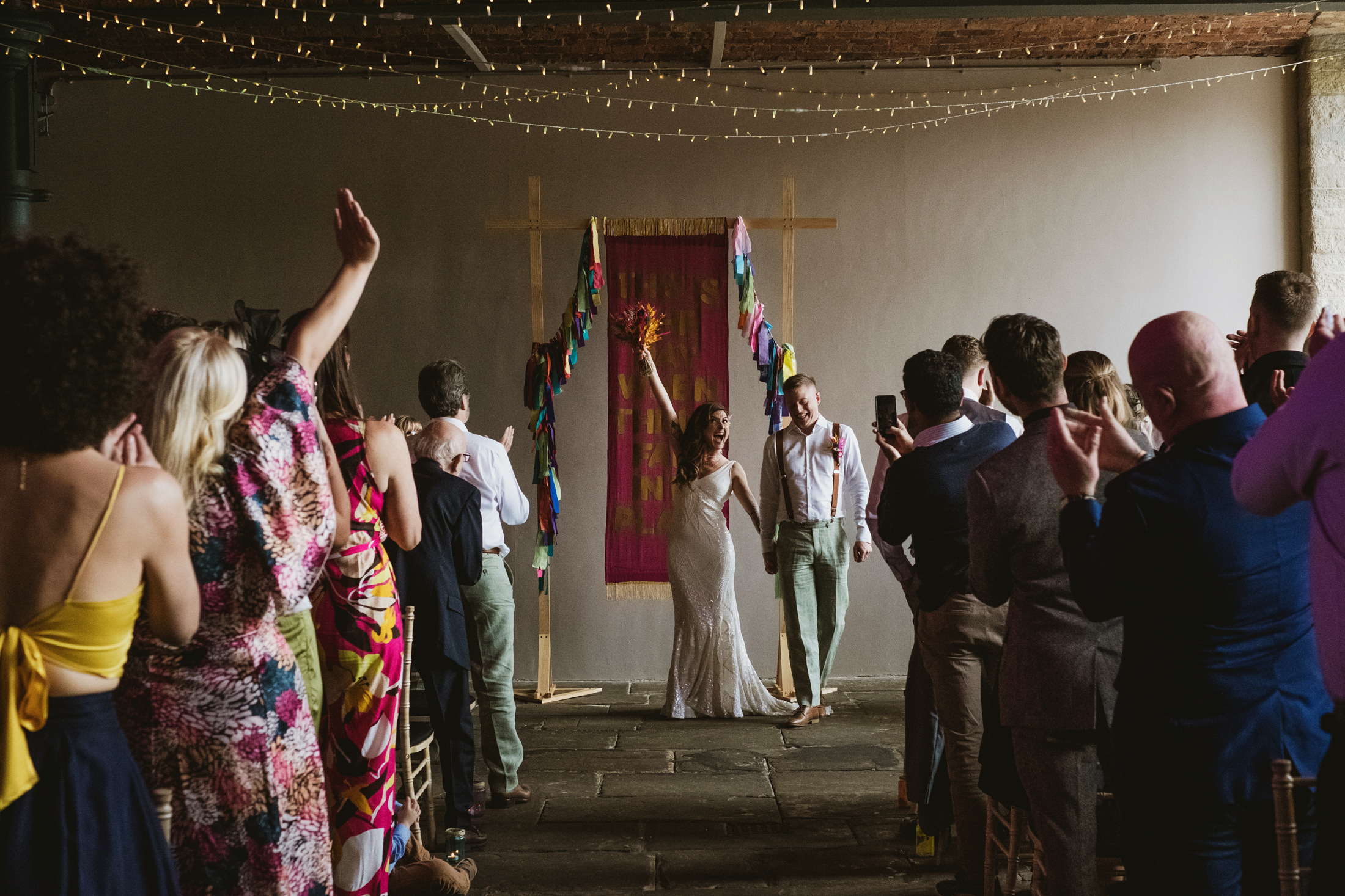 Bride and groom raise hands, laughing beneath fairy lights as guests applaud during documentary wedding photography.