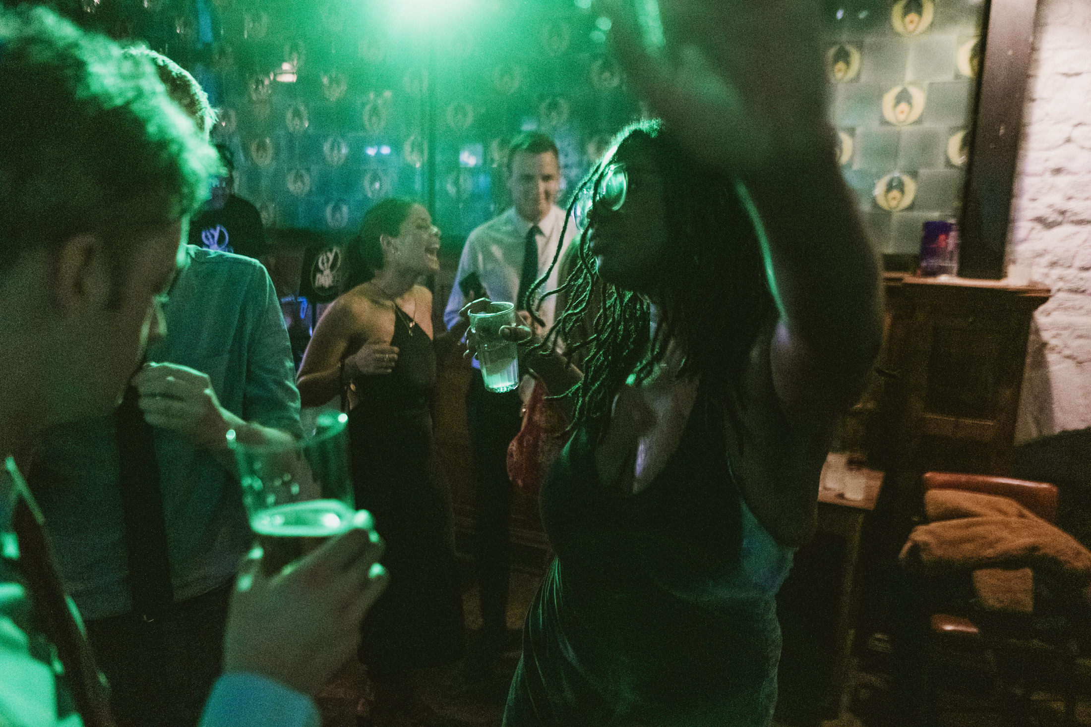 Guests laughing and dancing with drinks beneath green lights at a lively wedding reception