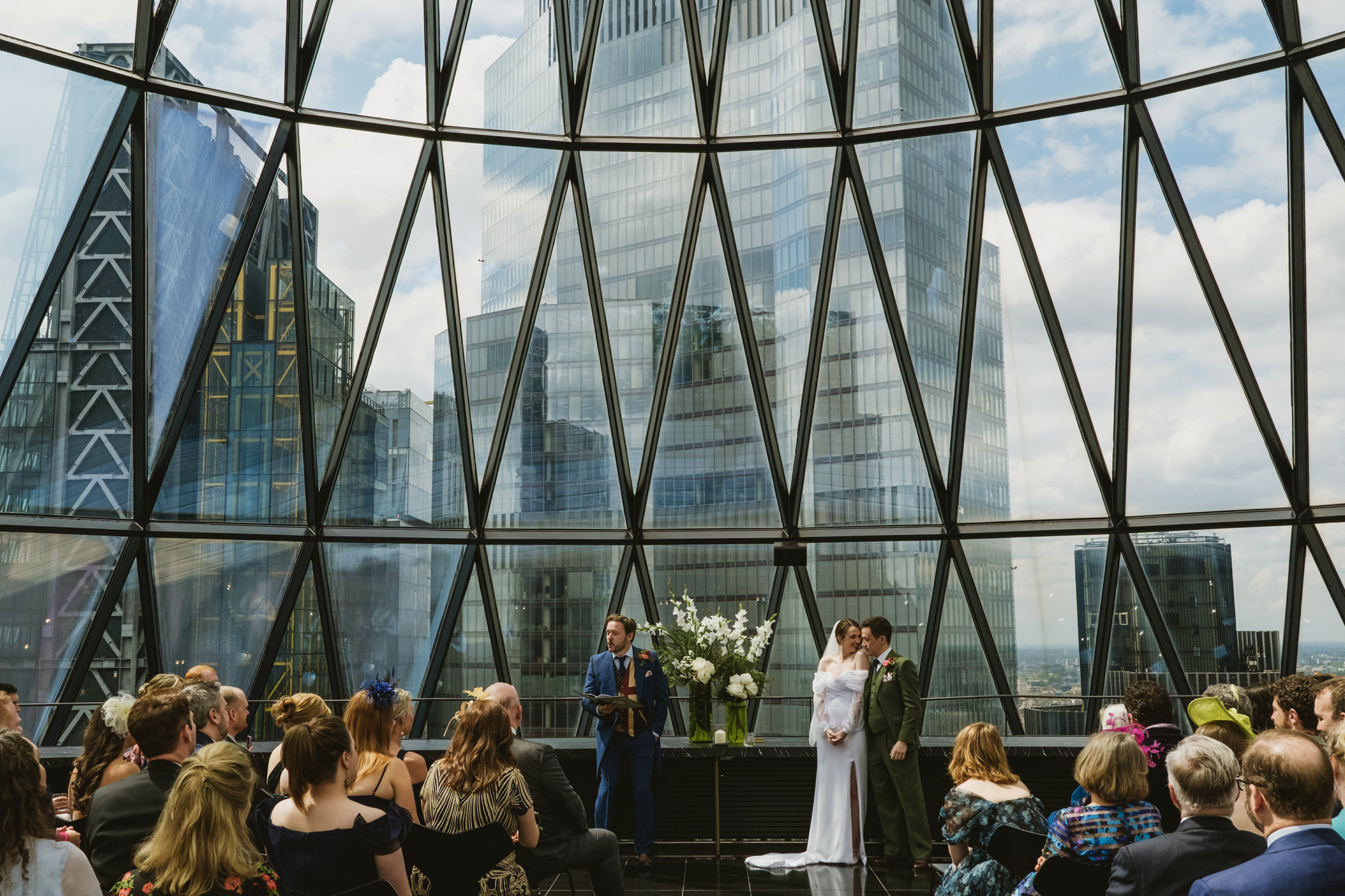 Bride and groom stand beside officiant during city wedding ceremony beneath vast glass windows.