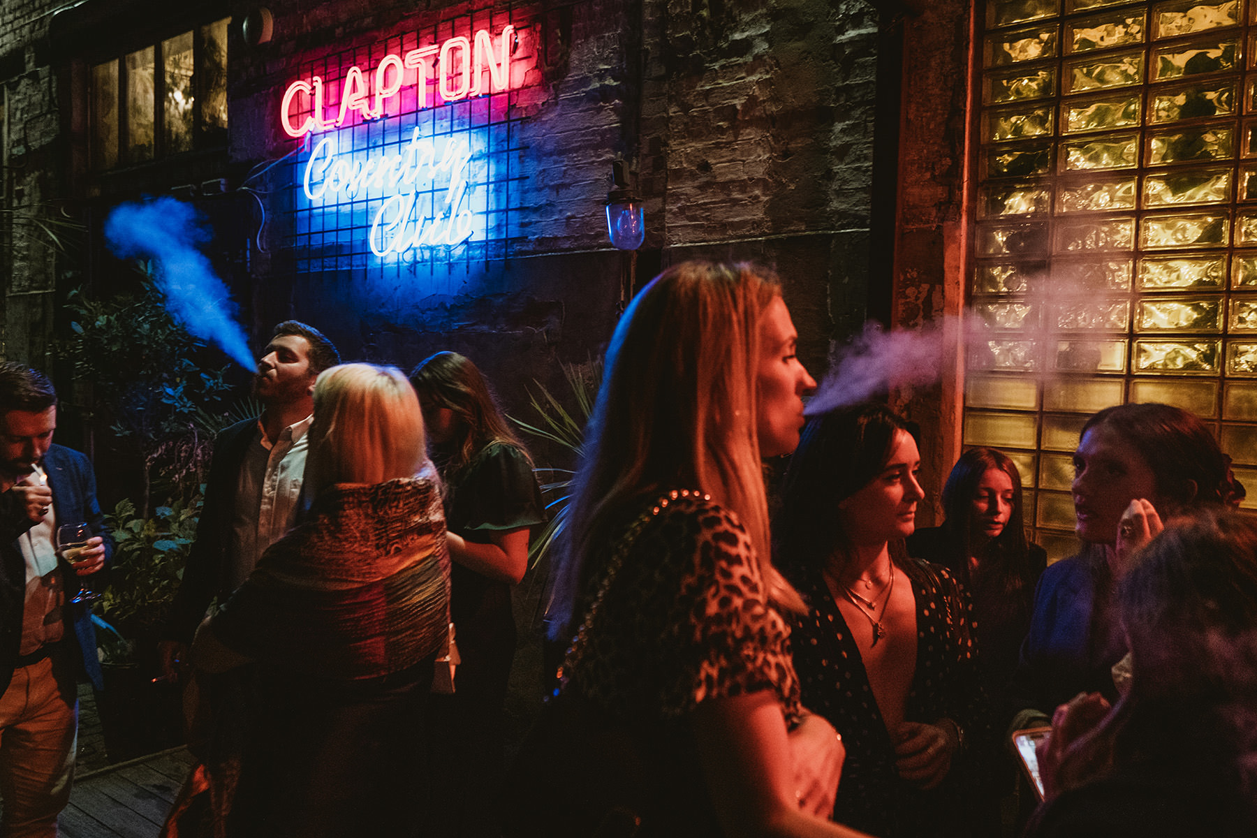 A candid, night-time documentary photograph outside Clapton Country Club featuring guests drinking and laughing under the glow of the venue's iconic neon signage.